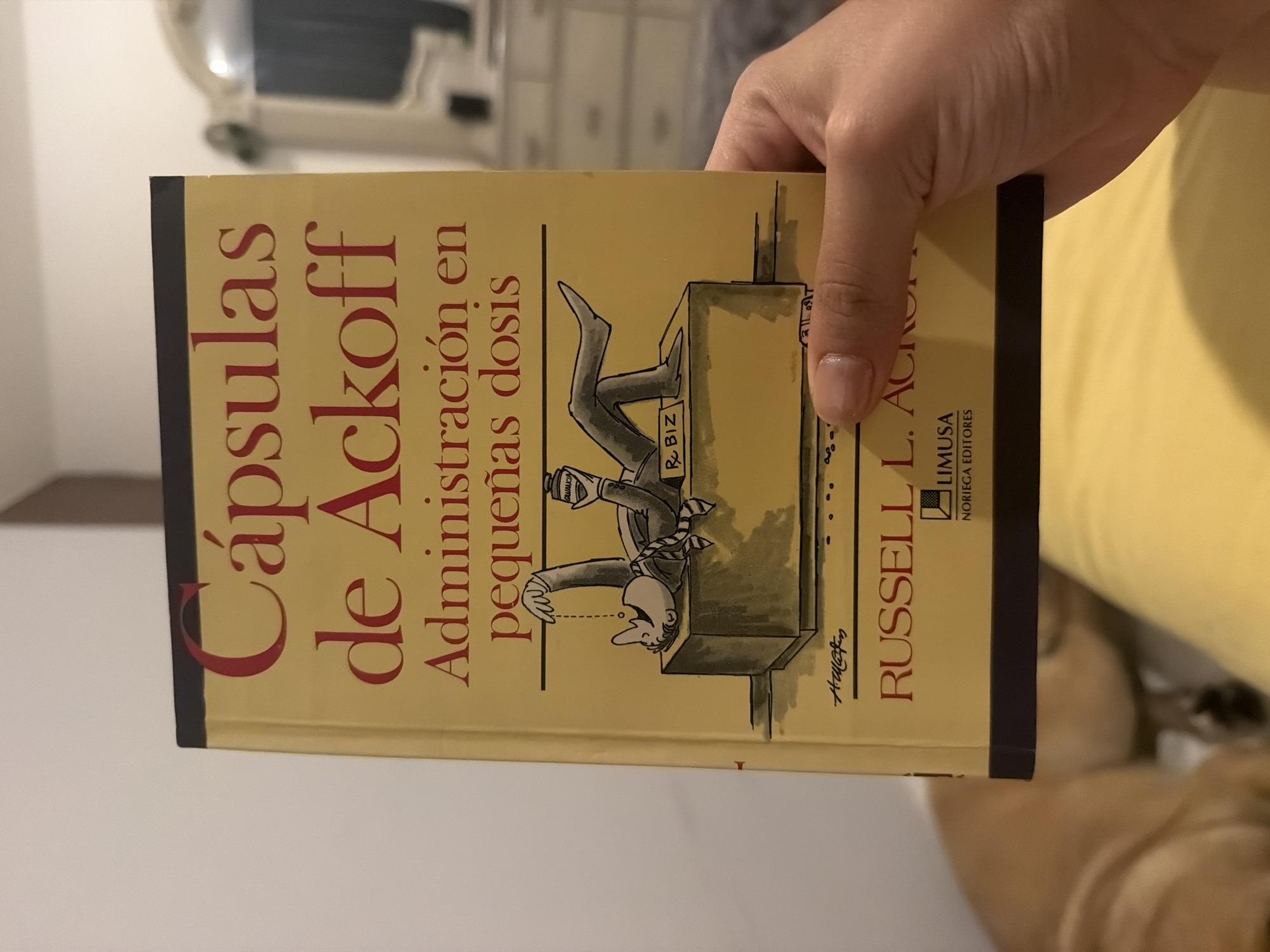 A hand holds a book titled "Cápsulas de Ackoff, Administración en pequeñas dosis" by Russell L. Ackoff. The book's cover has a cartoon drawing of a man upside down, his feet sticking out from under a desk labeled "R. Biz". He is drinking from a bottle labeled "Ganancia". The publisher is Limusa Noriega Editores. The location appears to be indoors, potentially a home or office, as indicated by the furniture and other objects in the background. There's no specific evidence to indicate this is in Santa Clara Ocoyucan, Mexico.