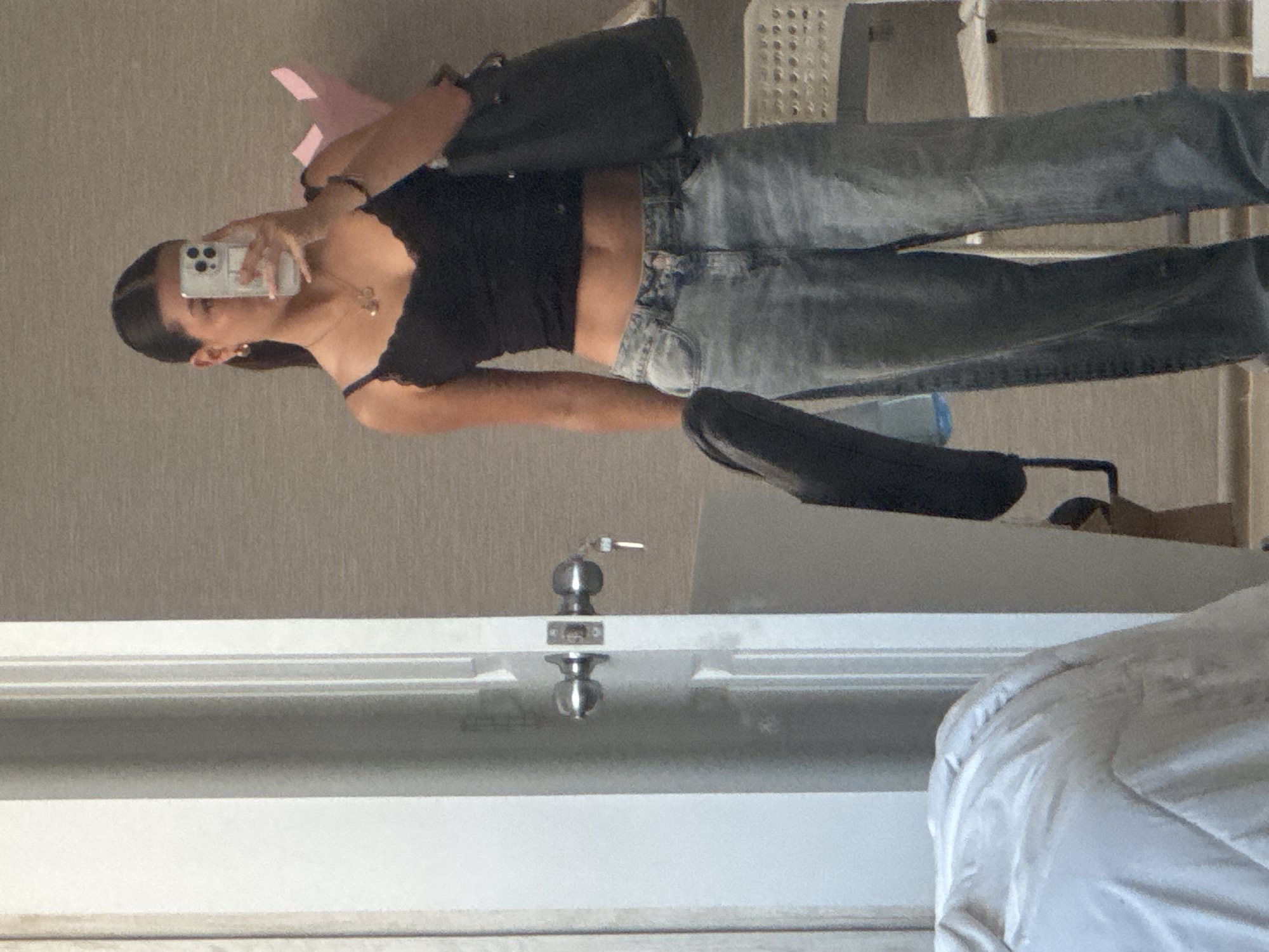 A person is taking a mirror selfie indoors. The person has dark hair pulled back, is wearing a black top, and jeans. They are holding a smartphone in their right hand. A black purse is draped over their left shoulder. The background is a gray wall. A white chair is visible in the upper right corner. A partially visible white bed is in the bottom right corner. A door with silver knobs is visible in the foreground. The location is not identifiable from the image.
