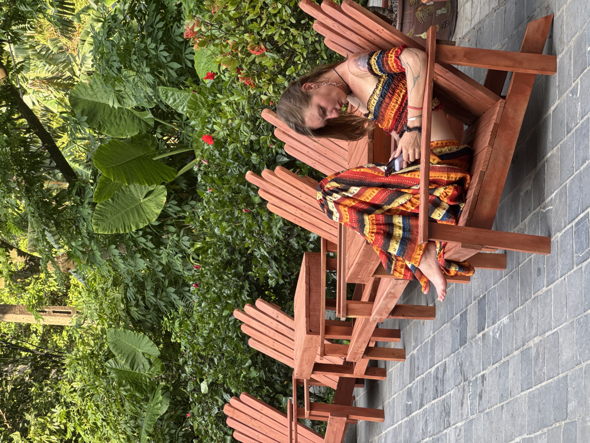 A woman sits in a red Adirondack chair, looking at her phone. She wears a colorful, off-the-shoulder top and a matching skirt with a striped pattern of yellow, red, orange, and black. Her feet are bare. The chair is made of wood and is one of several arranged in a row on a paved patio. Behind the chairs is a lush green garden with large-leafed plants and some red flowers. The scene appears to be outdoors, possibly in a resort or park setting, in Nam Hoa Lư Ward, Vietnam.