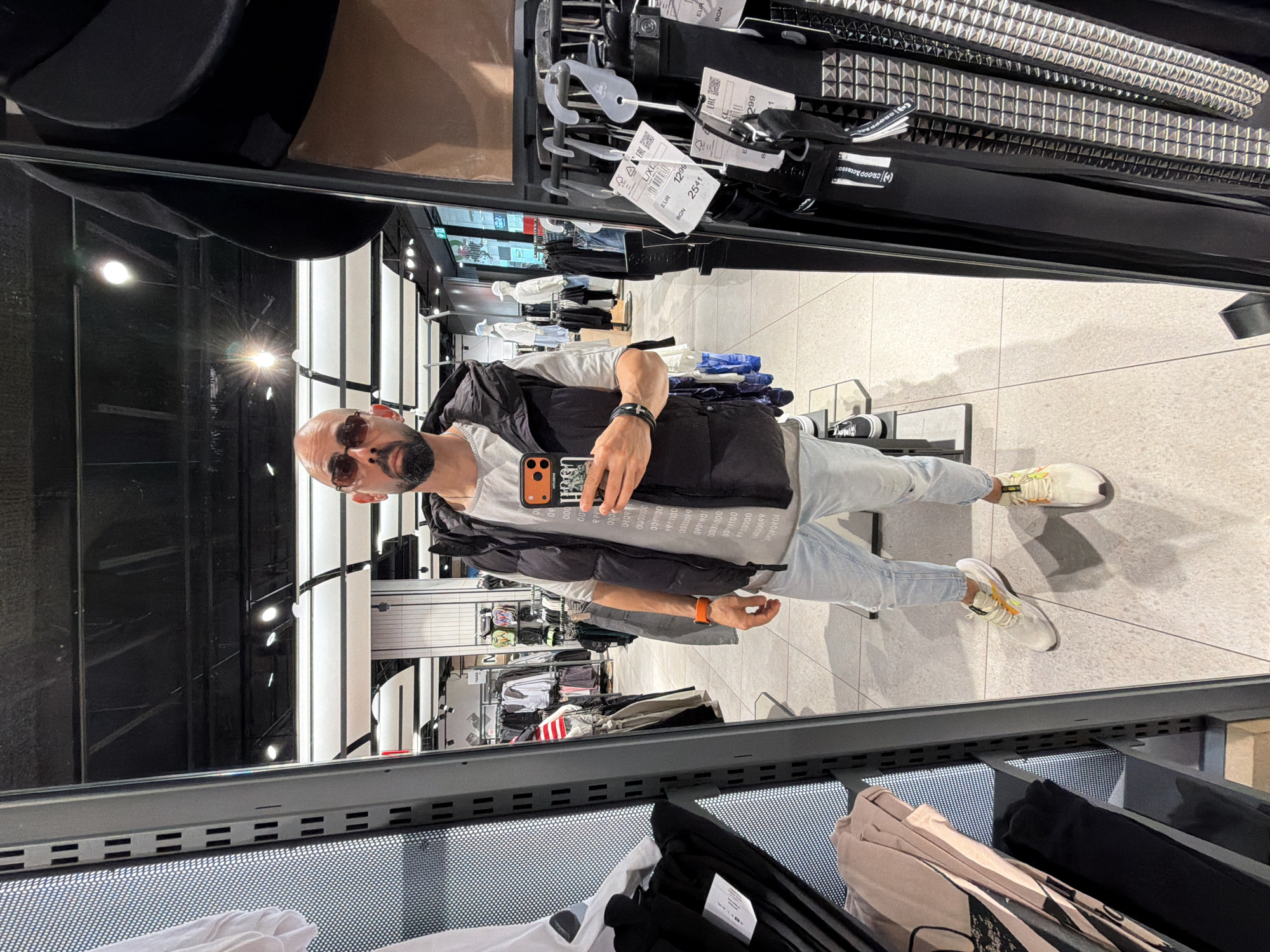 A full-length shot of a man in a mirror in a clothing store, possibly in Plovdiv, Bulgaria based on the OCR text. The man is wearing sunglasses, a white t-shirt with a pattern, a black vest, light-wash jeans, and white sneakers with orange accents. He is holding a cell phone with a patterned case. The reflection shows the interior of the store, with clothing racks and lighting fixtures visible. In the foreground, clothes are arranged in dark grey metal shelves. The OCR text indicates prices in EUR and BGN, which are currencies used in Bulgaria.