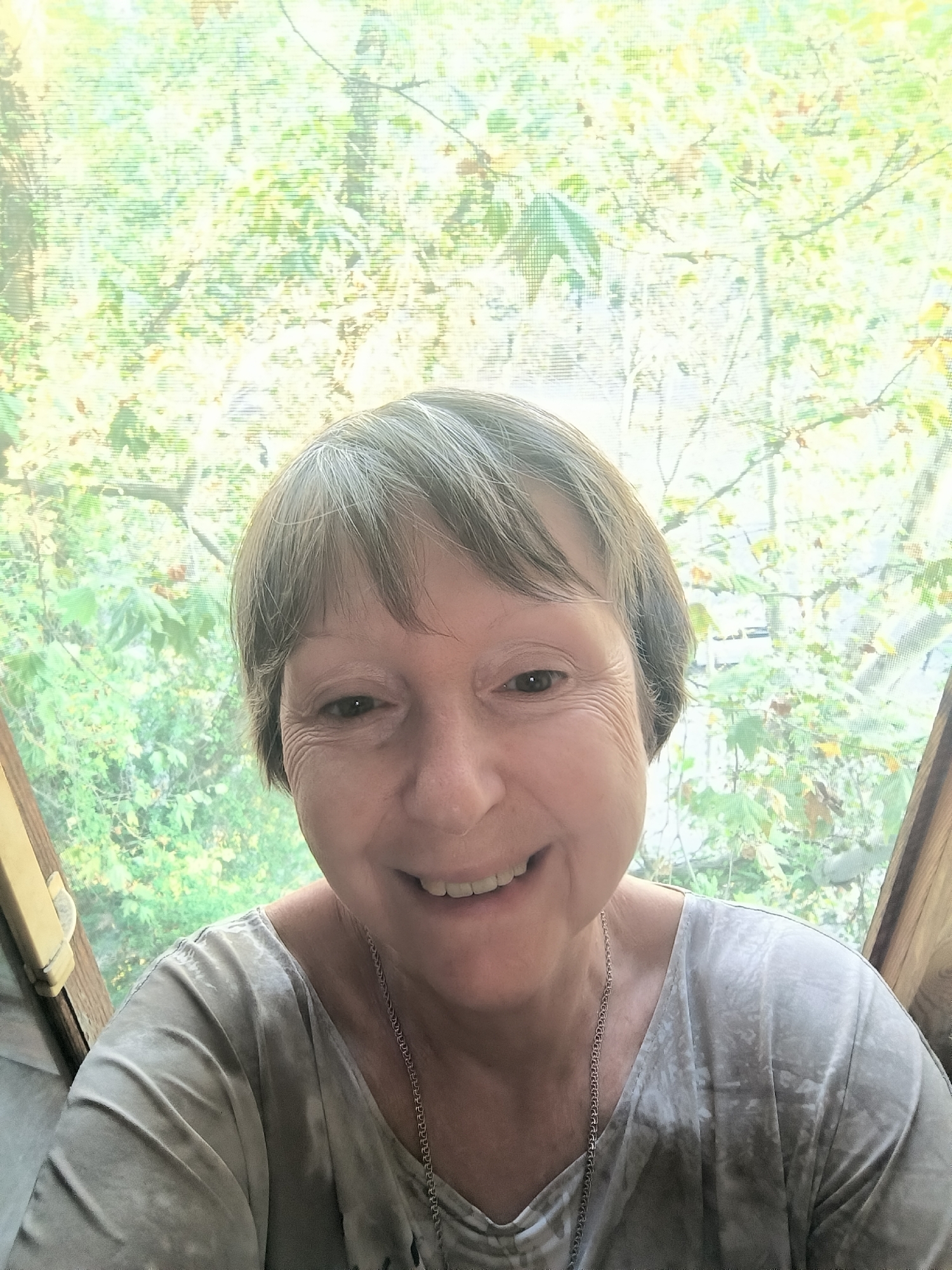 An outdoor selfie features a smiling Caucasian woman with short, light brown hair and fair skin, positioned centrally against a blurred background of green and yellow foliage, possibly indicating trees or bushes. She is wearing a light gray t-shirt with a tie-dye pattern and a silver chain necklace. The image appears to be taken during daylight, with natural light filtering through the foliage. The woman's face is the primary focal point. The setting does not contain any discernible landmarks or elements that would allow for identification of a specific location within Mykolaiv, Ukraine, or any other city. The image is a close-up, likely taken with a mobile device.