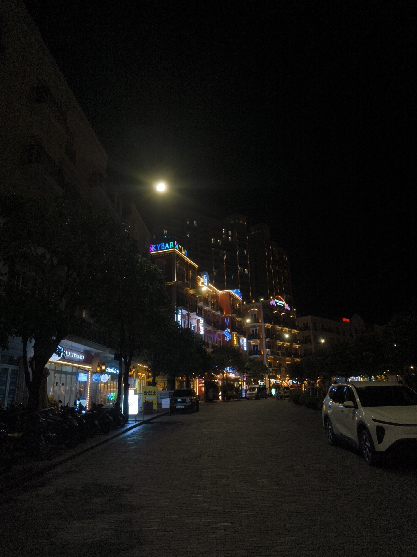 The image depicts a nighttime street scene in Phú Quốc, Vietnam. The street is paved with cobblestones and lined with trees on the left. On the left side of the street, there are several businesses with illuminated signs, including "BEAUTY STUDIO" and "Coco's". A row of parked motorcycles is visible in front of these businesses.
Across the street, a multi-story building is adorned with bright, colorful lights, including illuminated signs for "SKYBAR" and "BUFFET". The building has several balconies and windows, some of which are lit. Further down the street, other illuminated buildings can be seen.
A white SUV is parked on the right side of the street, facing away from the camera. The sky is dark, with a bright full moon visible above the buildings. The overall atmosphere suggests a lively urban environment at night.