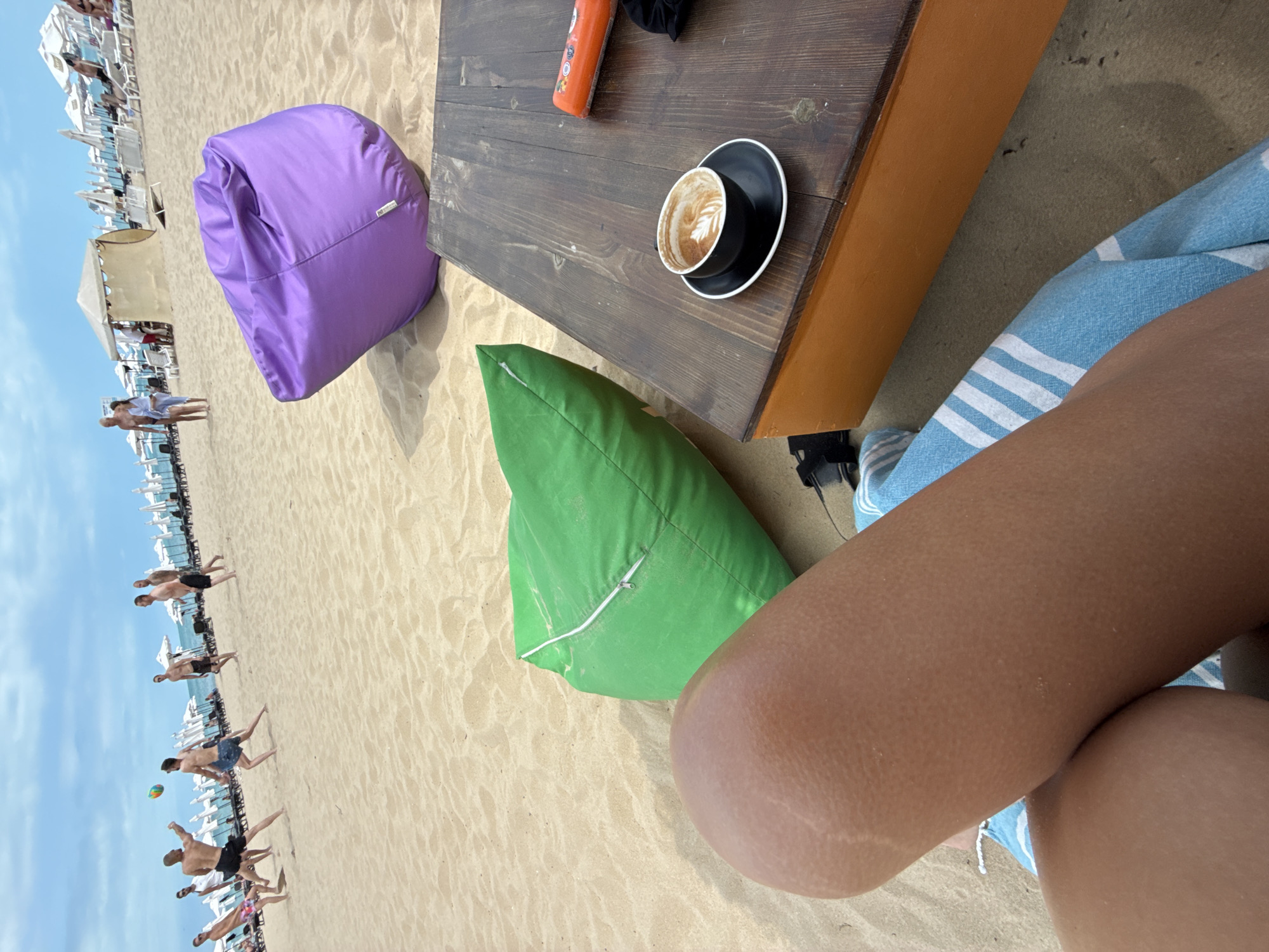 On a sandy beach in Nesebar, Bulgaria, several men are playing volleyball. In the foreground, a person's legs are visible, resting on a blue and white striped towel. Next to the legs is a large, green beanbag chair. Further into the scene, a purple beanbag chair sits on the sand. A wooden table is positioned to the right, with a cup of coffee and an orange object on it. In the background, beach chairs and umbrellas are set up, and more people are visible, some walking and others relaxing. The sky is partly cloudy.