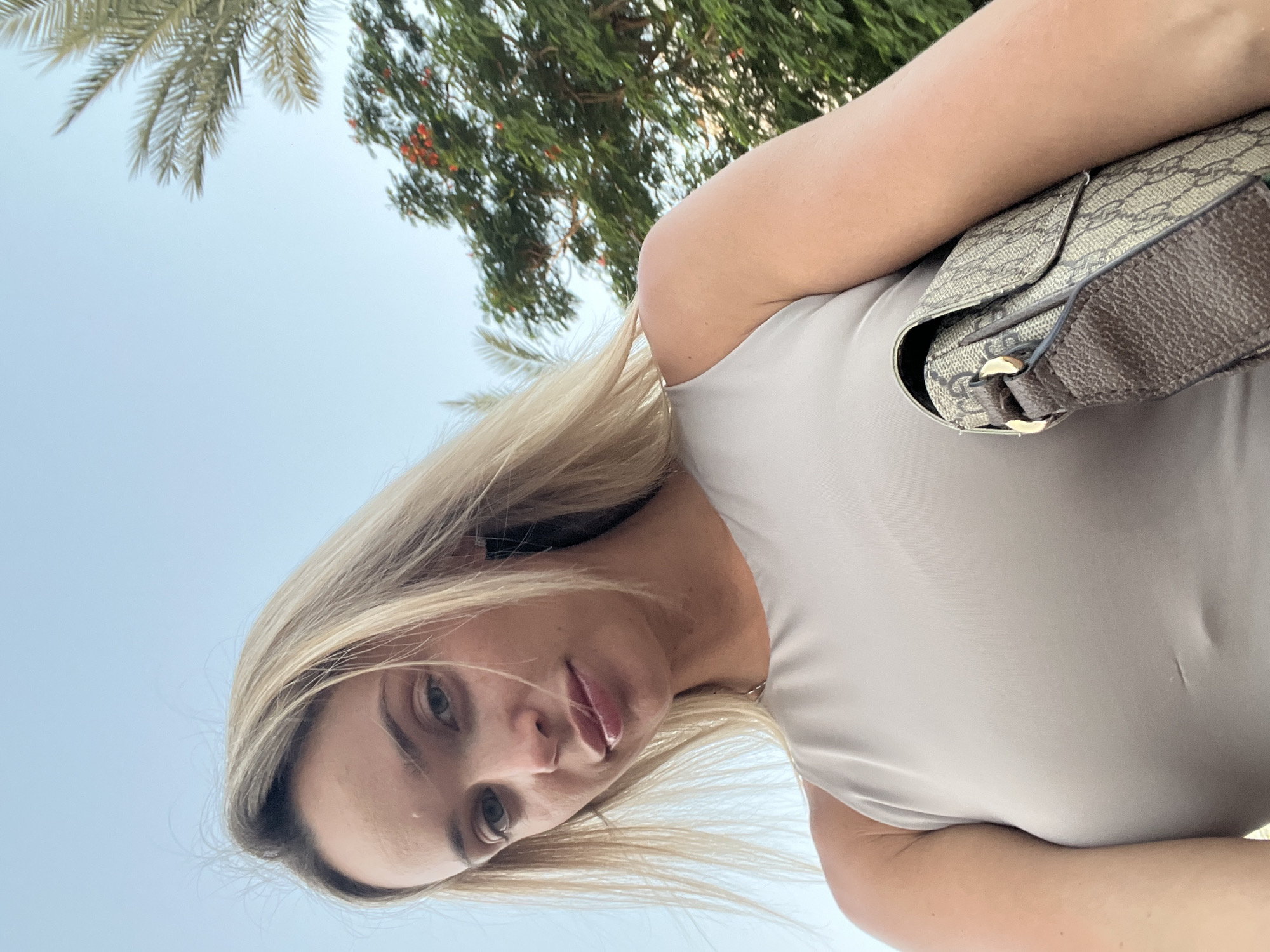 A young woman with blonde hair and light skin is looking down at the camera. She is wearing a beige tank top and holding a beige and brown patterned purse. The background shows a bright blue sky with some palm tree fronds and green foliage with red flowers. The image appears to be taken outdoors in a warm climate, possibly a garden or park area in Sharm El Sheikh, Egypt, as suggested by the palm trees and lush vegetation.