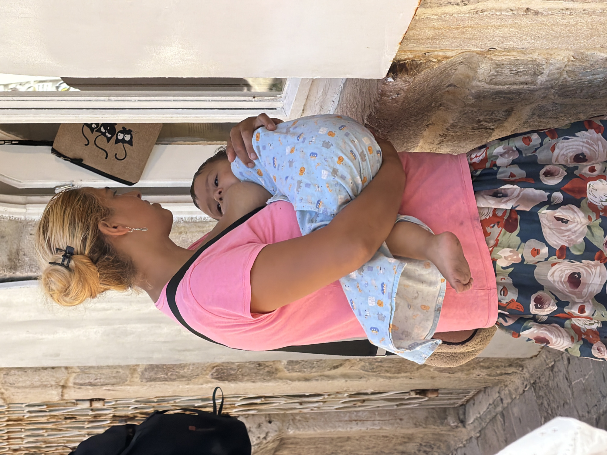 A woman with blonde hair tied up in a bun, wearing a bright pink t-shirt with black trim, holds a baby wrapped in a blue onesie with a cartoon pattern. The baby is facing the viewer, with its head resting on the woman's shoulder. The woman is looking at the baby with a tender expression. She is wearing a floral skirt with large pink and white roses.
In the background, there is a textured stone wall and a white surface which could be a wall or a ledge. Below the woman, a black backpack is visible, resting on a stone-tiled surface. A small, brown pouch with a black cat print is tucked behind the woman's head, against the white surface. The scene appears to be outdoors, possibly in a courtyard or alleyway, with the lighting suggesting it is daytime. The overall mood is one of care and affection. There is no visible text, other than the cat print on the pouch. The setting of Budva, Montenegro, with its characteristic stone architecture, is consistent with the visual elements.