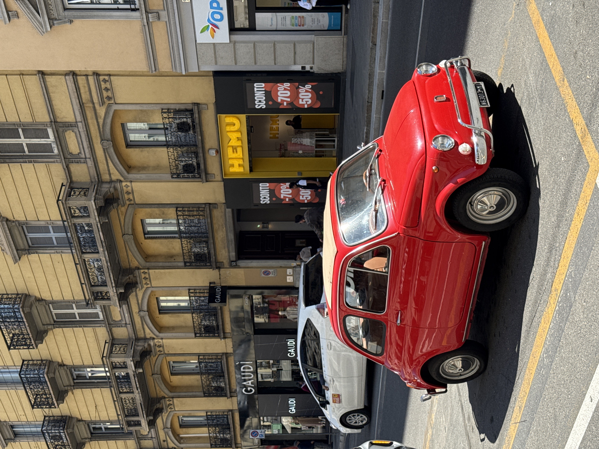 A red vintage Fiat 500 is parked on a street in Milan, Italy, next to a pedestrian crossing with yellow markings. The car has an Italian license plate, N63013-MI. The street features stores such as "HEMU" and "GAUDI," with yellow and black storefronts. Building facades are visible with ornate iron balconies and cream-colored walls. The lighting suggests a sunny day. A modern white car is partially visible behind the Fiat.