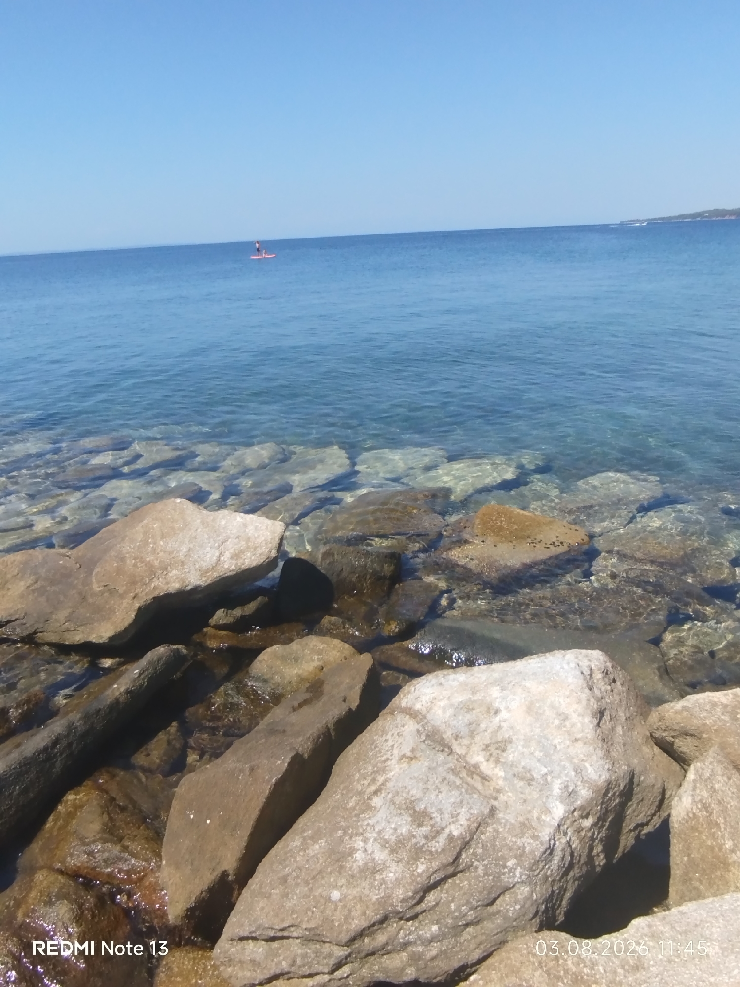The image depicts a clear, blue sky over a calm, transparent sea. In the distance, a person is visible on a paddleboard in the water. The foreground is dominated by large, textured rocks, some partially submerged in the shallow, clear water, allowing the seabed stones to be seen. The lower left corner of the image displays the text "REDMI Note 13," and the lower right corner shows the date and time "03.08.2026 11:45." The scene suggests a peaceful coastal environment, likely in Sithonia, Greece, given the context provided.