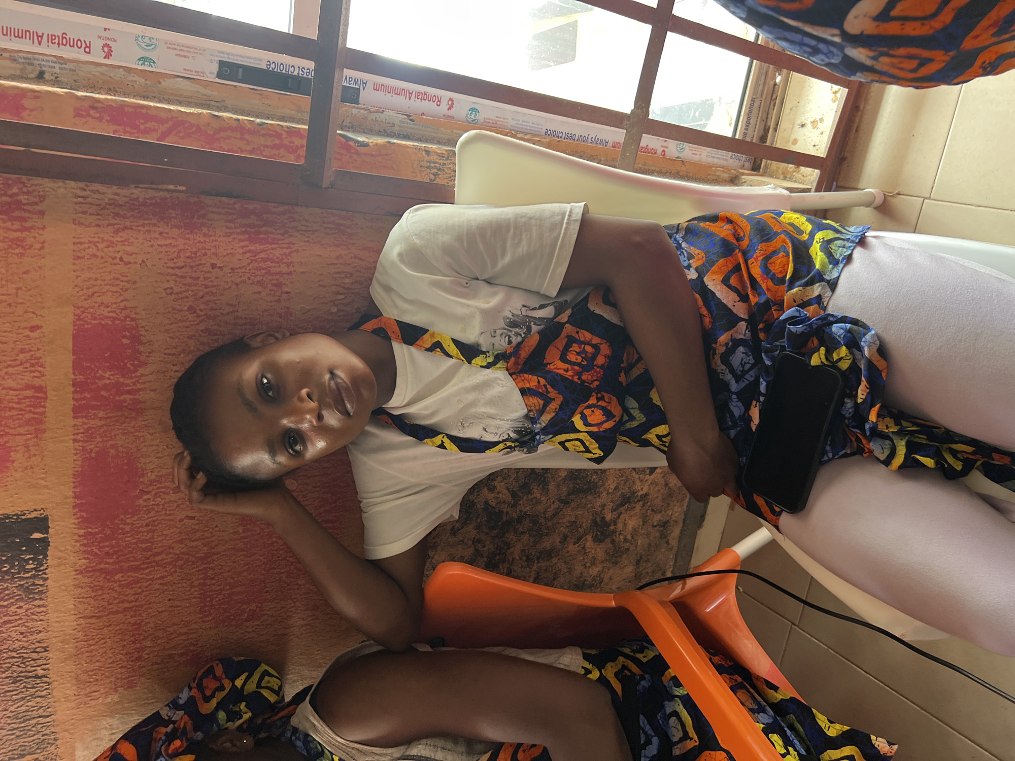 A young woman is lying down, propped up on her elbow, looking at the camera. She is wearing a white t-shirt and a colorful, patterned fabric around her waist. Her legs are extended in front of her. A black smartphone is visible on her lap, partially covered by the fabric. Two bright orange plastic chairs are in the foreground. The background shows a window with some text visible on the window frame, which includes "Rongtai Aluminium Alway" and "best choice". The overall setting appears to be indoors, possibly a waiting area or a common space, with a tiled wall and a textured orange wall in the background. The lighting suggests it is daytime. The woman's expression is neutral, possibly relaxed or contemplative.