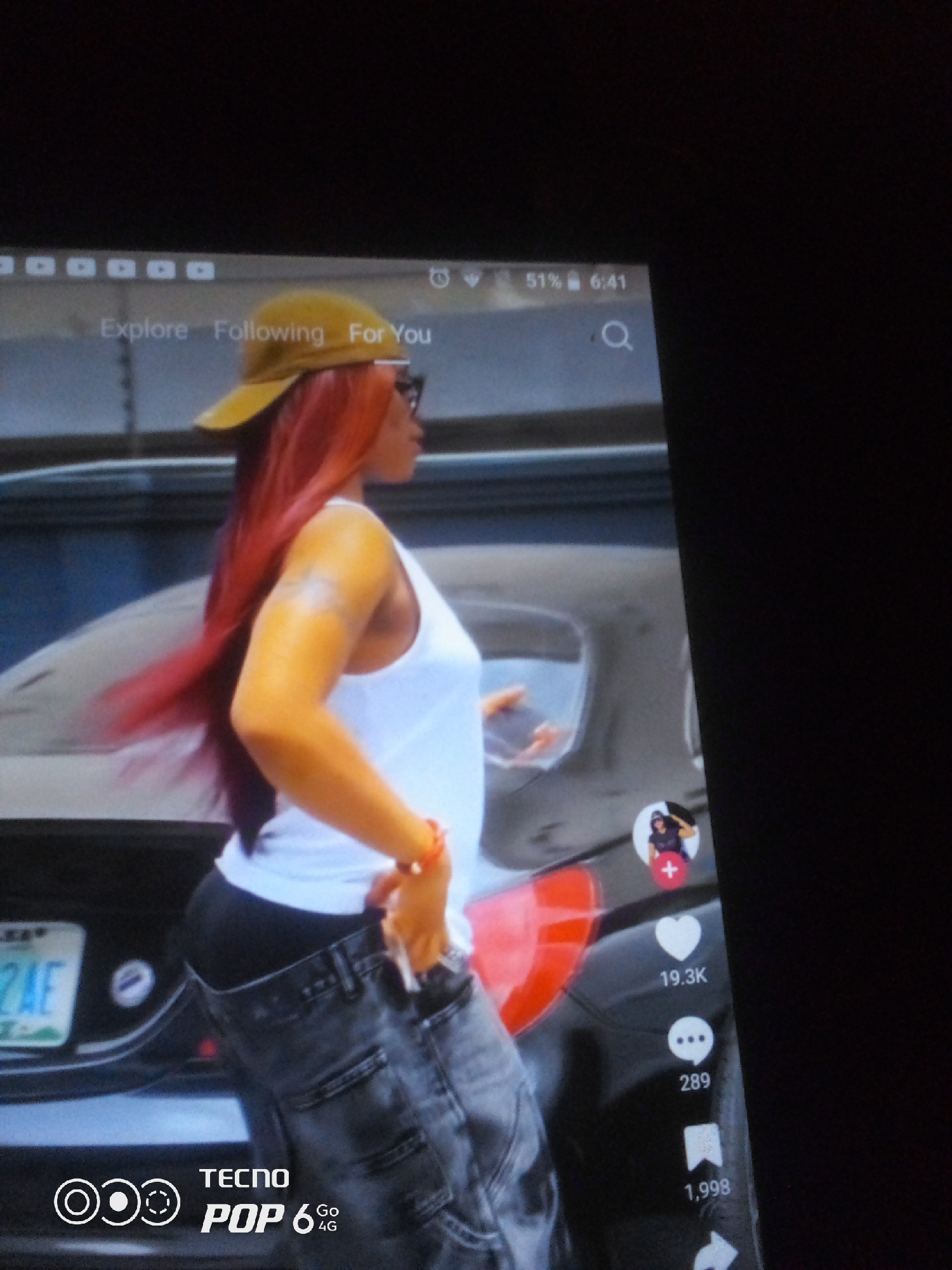 This is a full-screen, slightly angled, close-up, indoor, digital photograph of a woman with long, bright red hair wearing a yellow baseball cap, a white tank top, and loose-fitting, dark-colored jeans. She is looking to her right, with her right arm bent at the elbow and her hand near her waist. She has a bright orange bracelet on her right wrist. The woman is standing next to a dark-colored vehicle. A license plate with the characters "2AE" is visible on the rear of the vehicle. The background is dark and out of focus. The image appears to be a screenshot from a mobile device, as evidenced by the on-screen interface elements, including a search bar, social media icons, engagement metrics (19.3K likes, 289 comments, 1,998 shares), and branding for "TECNO POP 6 4G". There is no indication of the specific location within Howayi, Nigeria, or any other geographical details. The lighting is dim, with the primary light source appearing to come from the device screen.
