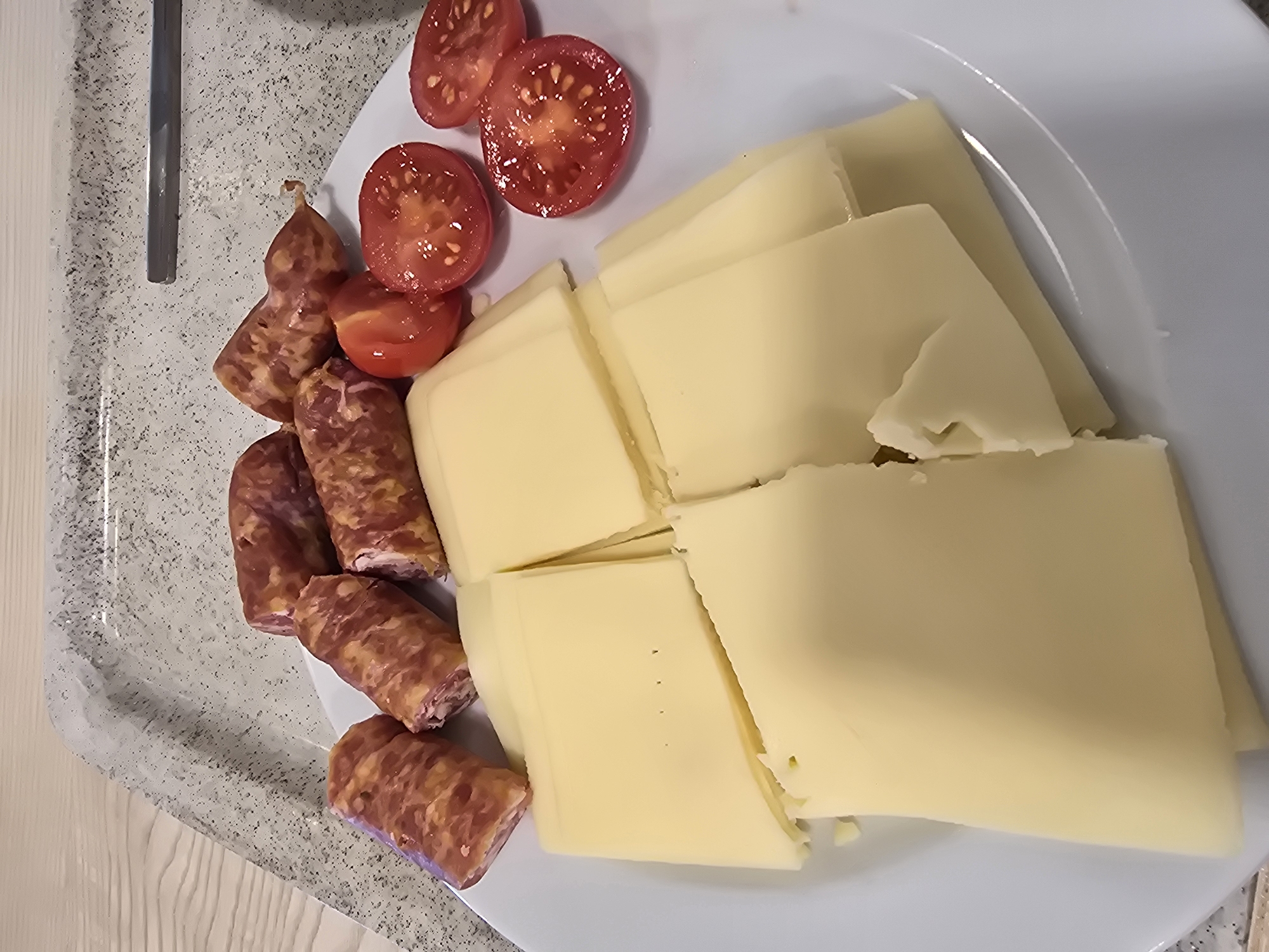A white plate containing sliced cheese, sliced tomatoes, and sliced sausages is on a speckled gray tray. The tray is on a light-colored wooden table. The background shows a portion of a gray countertop with a metallic object on it. The image appears to be a food presentation, possibly at a restaurant or a catered event in Garmisch-Partenkirchen, Germany, due to the presence of local sausages and cheese, common in Bavarian cuisine. The lighting is even, suggesting an indoor setting. There are no people or animals visible in the image.