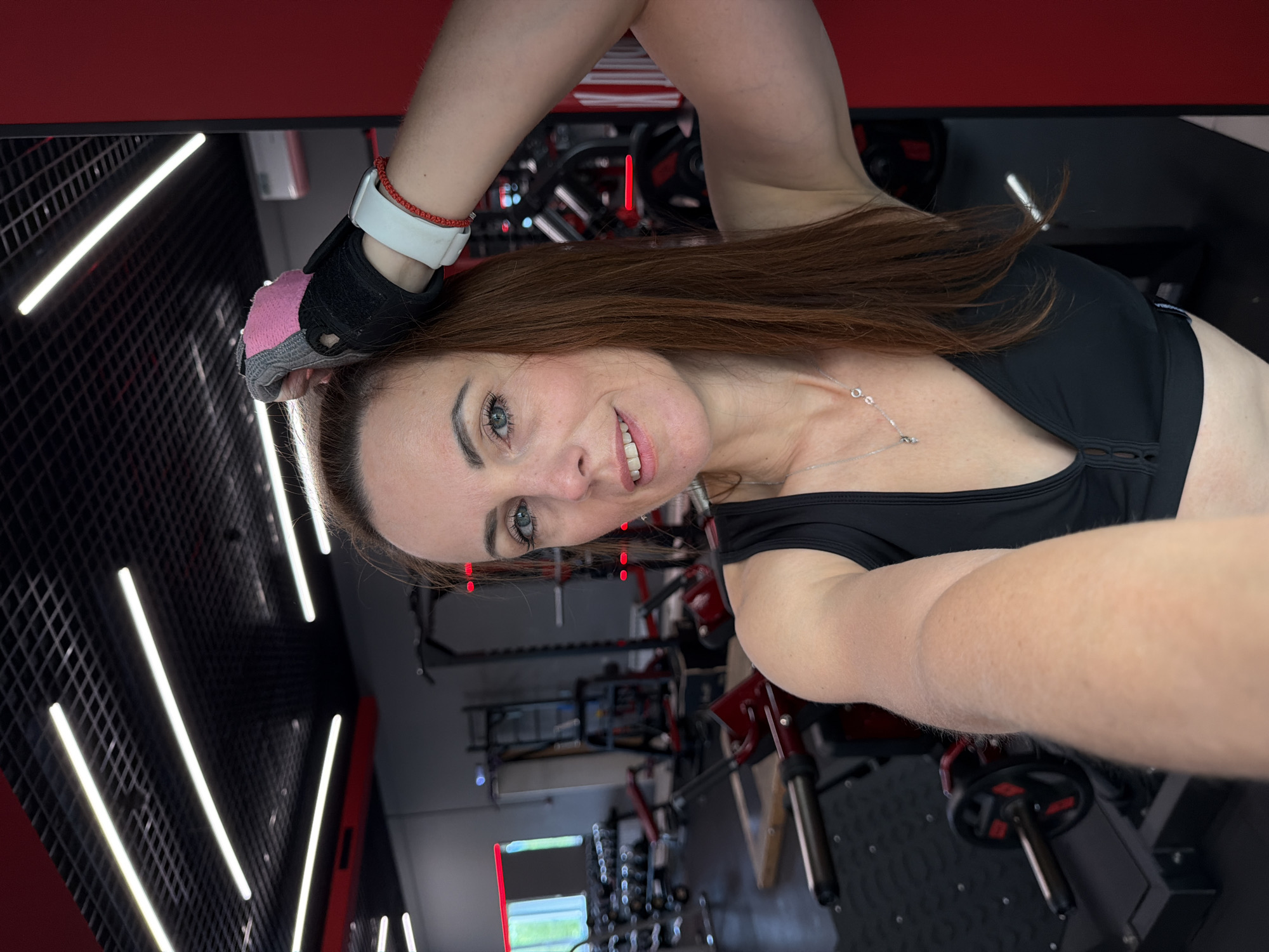 A woman with brown hair and blue eyes, wearing a black sports bra, a workout glove on her right hand, and a white wristband with a red string bracelet, poses for a selfie in a gym. The gym has a red and black color scheme with gym equipment visible in the background. The woman is looking at the camera and smiling. The location is not specified to be in Birobidzhan, Russia.