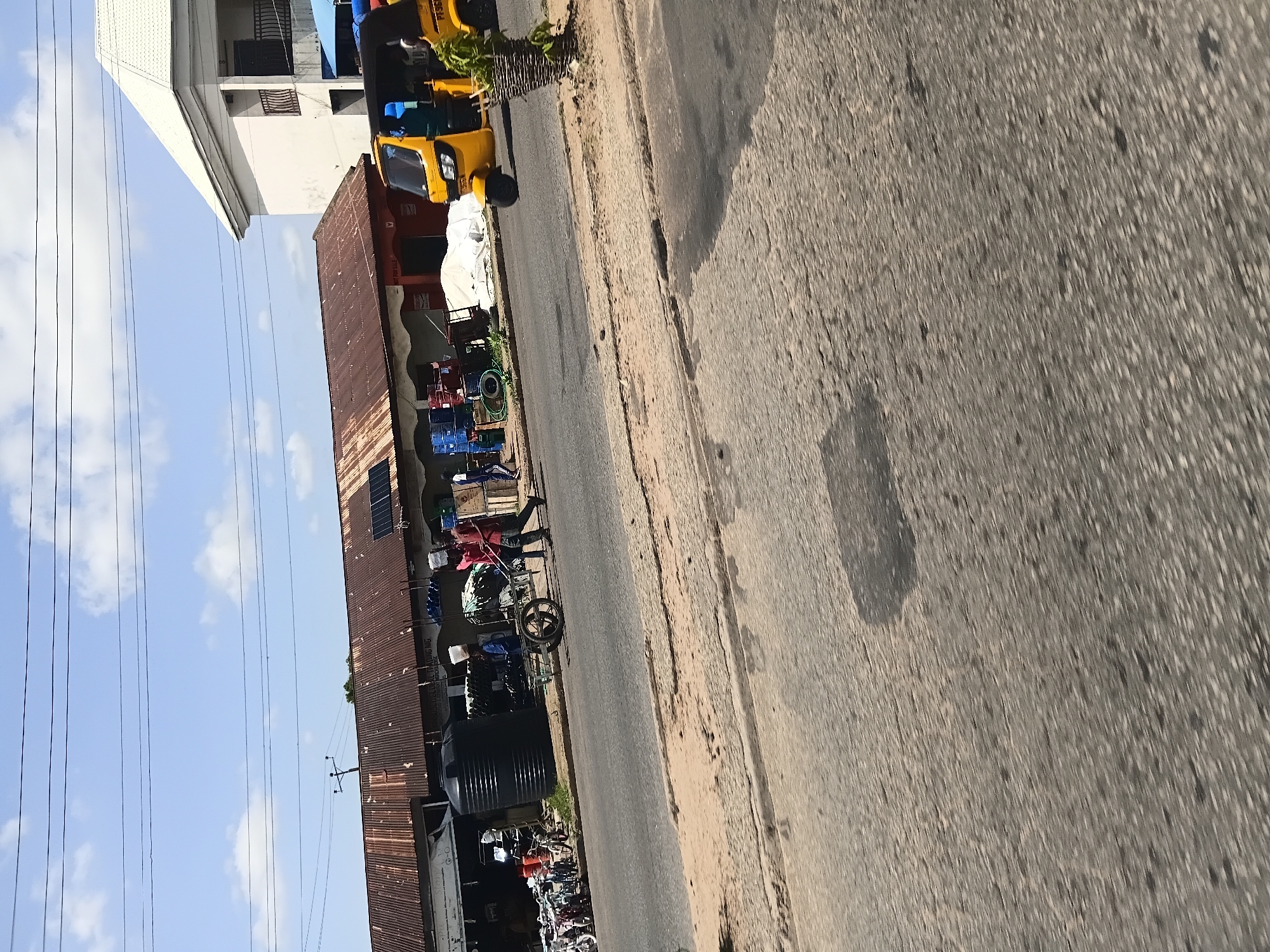 The image captures a bustling street scene in Jalingo, Nigeria, under a bright, partly cloudy sky. The foreground is dominated by a paved road with a sandy shoulder. In the mid-ground, a row of market stalls with corrugated iron roofs line the street, displaying a variety of goods. To the left, bicycles are parked, and a large black water tank is visible. Several people are engaged in activities, including a man pushing a cart and others walking. A yellow auto-rickshaw, a common mode of transport in the region, is moving along the road. In the background, a modern white building stands out against the market structures, and power lines crisscross the sky. The overall impression is one of a lively commercial area with a blend of traditional and modern elements.