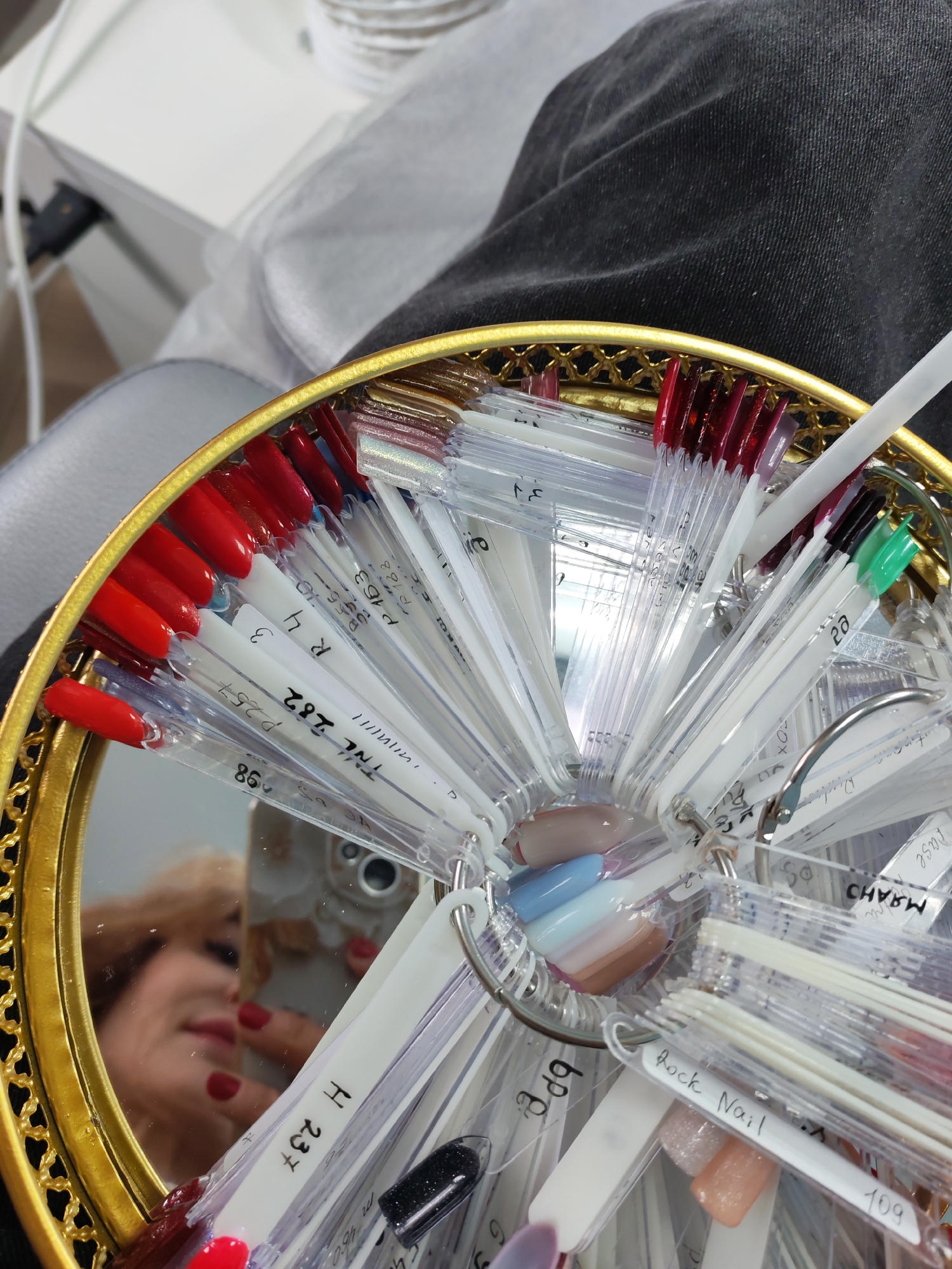 The image depicts a close-up view inside what appears to be a nail salon in Chaikovsky, Russia, focusing on a circular display of nail polish samples. The samples, presented on clear plastic sticks, are arranged on metal rings within an ornate, gold-framed mirrored tray.
Reflected in the mirror is the blurred but discernible face of a woman with light-colored hair, red lipstick, and red nail polish, holding a smartphone, suggesting the image is a selfie. She exhibits a subtle smile, indicating engagement or contentment while examining the polish options.
The nail polish collection showcases a variety of colors, with a significant number of red and dark red shades on the left, alongside pastel tones like light blue, pink, and beige, as well as glittery and black polishes. Many swatch sticks bear handwritten labels, including codes and numbers like "24", "P163", "P188", "R4", "P257", "TNL 282", "86", "109", and "H 237". Other visible text includes "aras", "Oi.", "D", "FOX", "Lase", "МЯАНО" (likely "MARANO"), and "Rock Nail". The background partially reveals a leg clad in dark gray denim and some white furniture or equipment, consistent with a professional beauty setting.