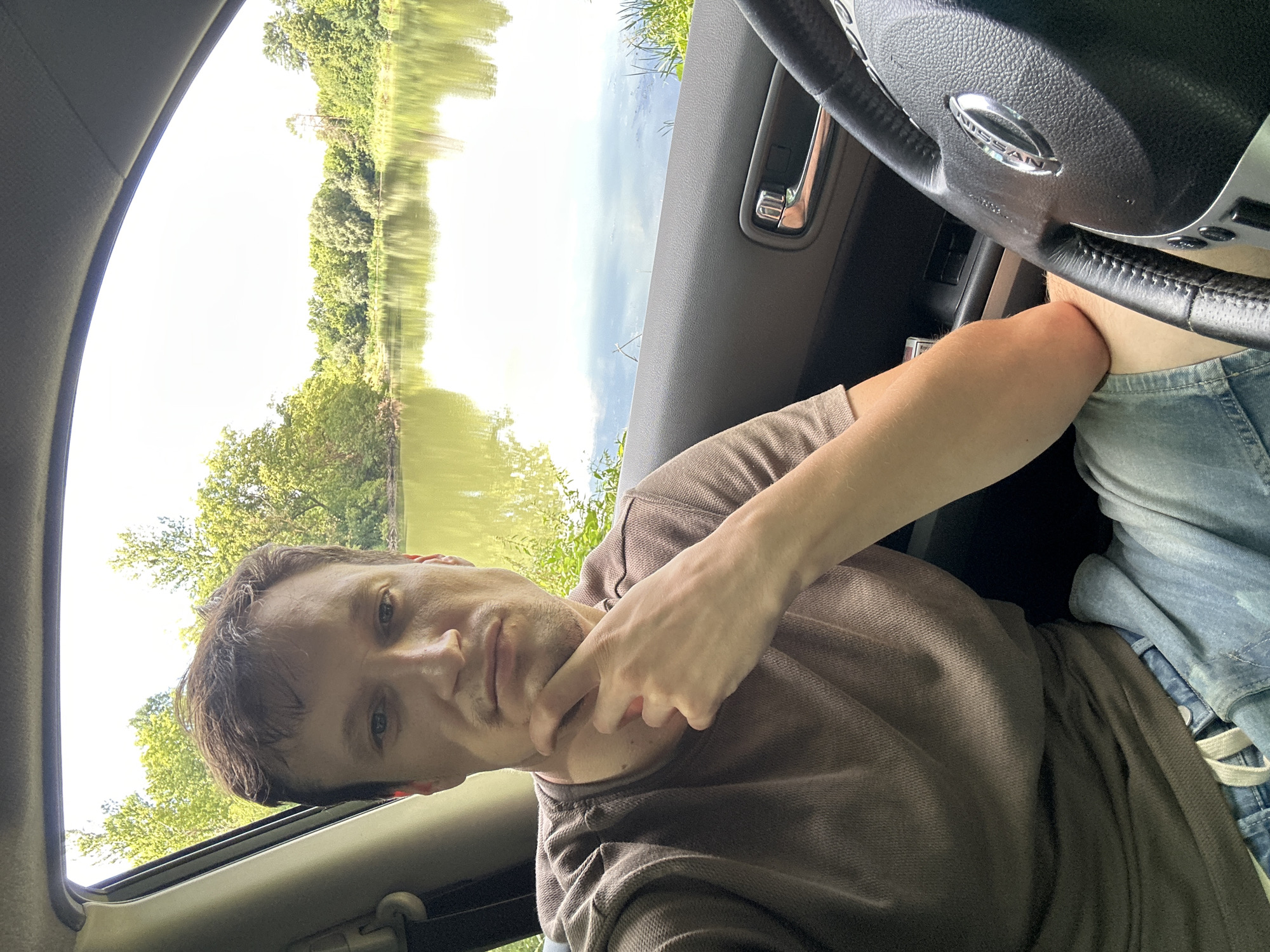 A man is sitting in the driver's seat of a car. He is looking at the camera with his chin resting on his hand. The car is parked next to a body of water, with trees and a bright sky visible in the background. The man is wearing a brown t-shirt and denim shorts. The steering wheel of the car is visible in the foreground. The scene appears to be a serene outdoor setting.