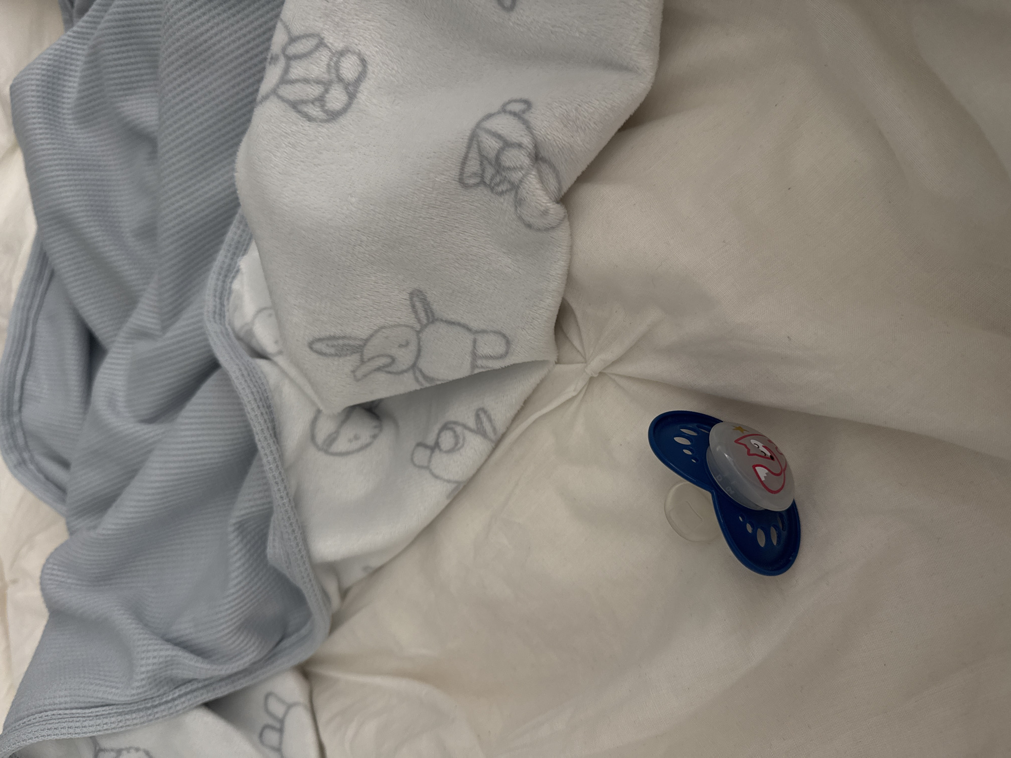 A blue and clear pacifier with a pink fox design lies on a white quilted surface. The pacifier is positioned at the bottom left of the image. Behind the pacifier, a light blue blanket with a ribbed texture is draped over a white blanket patterned with small gray drawings of animals, likely rabbits and bears. The scene appears to be indoors, on a bed or crib, in Siegburg, Germany.