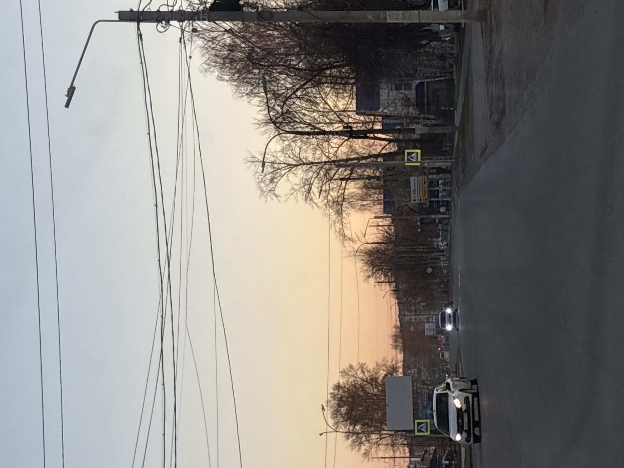 The image depicts a street scene in Chaikovsky, Russia, during dusk or dawn. The sky displays a gradient of colors ranging from light gray to orange-pink. Power lines stretch across the left side of the frame, connected to a utility pole topped with a street light. Trees line the side of the road, their bare branches silhouetted against the sky. The street is paved and appears to be a two-lane road with cars driving on it. A white vehicle is visible in the foreground, while another with headlights on is approaching in the distance. Pedestrian crossing signs are visible on the roadside. A billboard-like structure is also present near the bottom of the frame.