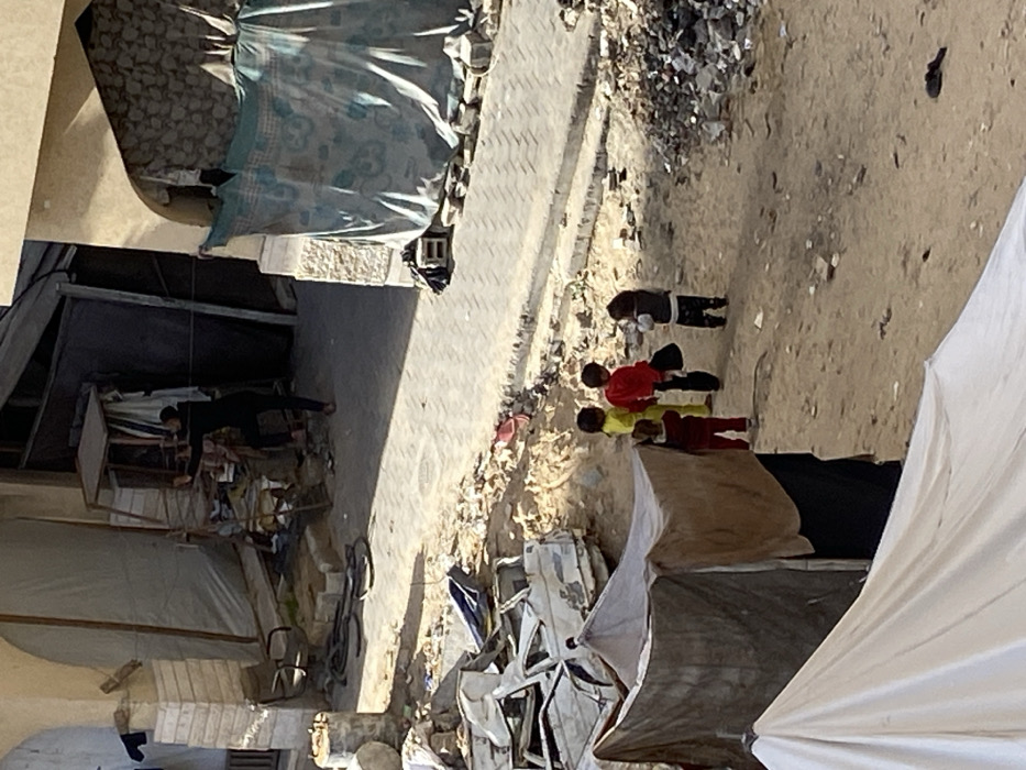 The image captures an outdoor scene in Khan Yunis, Palestinian Territory, characterized by urban debris and makeshift arrangements, viewed from an elevated position. In the upper-left, an adult individual in dark clothing stands near a rudimentary metal-framed structure or stall, adjacent to a building partially concealed by dark tarpaulins. To the right, another building entrance is covered by a blue patterned fabric, secured at its base. The ground features a section of light-colored paver blocks in the mid-ground transitioning to disturbed dirt and debris.
In the mid-ground left, multiple severely damaged and rusted vehicle bodies are piled amidst construction debris. Four young children are observed standing together on the dirt path in the mid-ground center-right. One child wears a bright yellow top, another a red top, a third a dark vest over patterned clothing while holding a dark object, and the fourth a dark red top and pants. The foreground left contains two makeshift shelters constructed from draped tarpaulins, one dark brown and another lighter brown, with a portion of a white tarp visible in the bottom-left. Scattered piles of rubble, dirt, and assorted waste materials are prevalent across the unpaved areas of the scene.