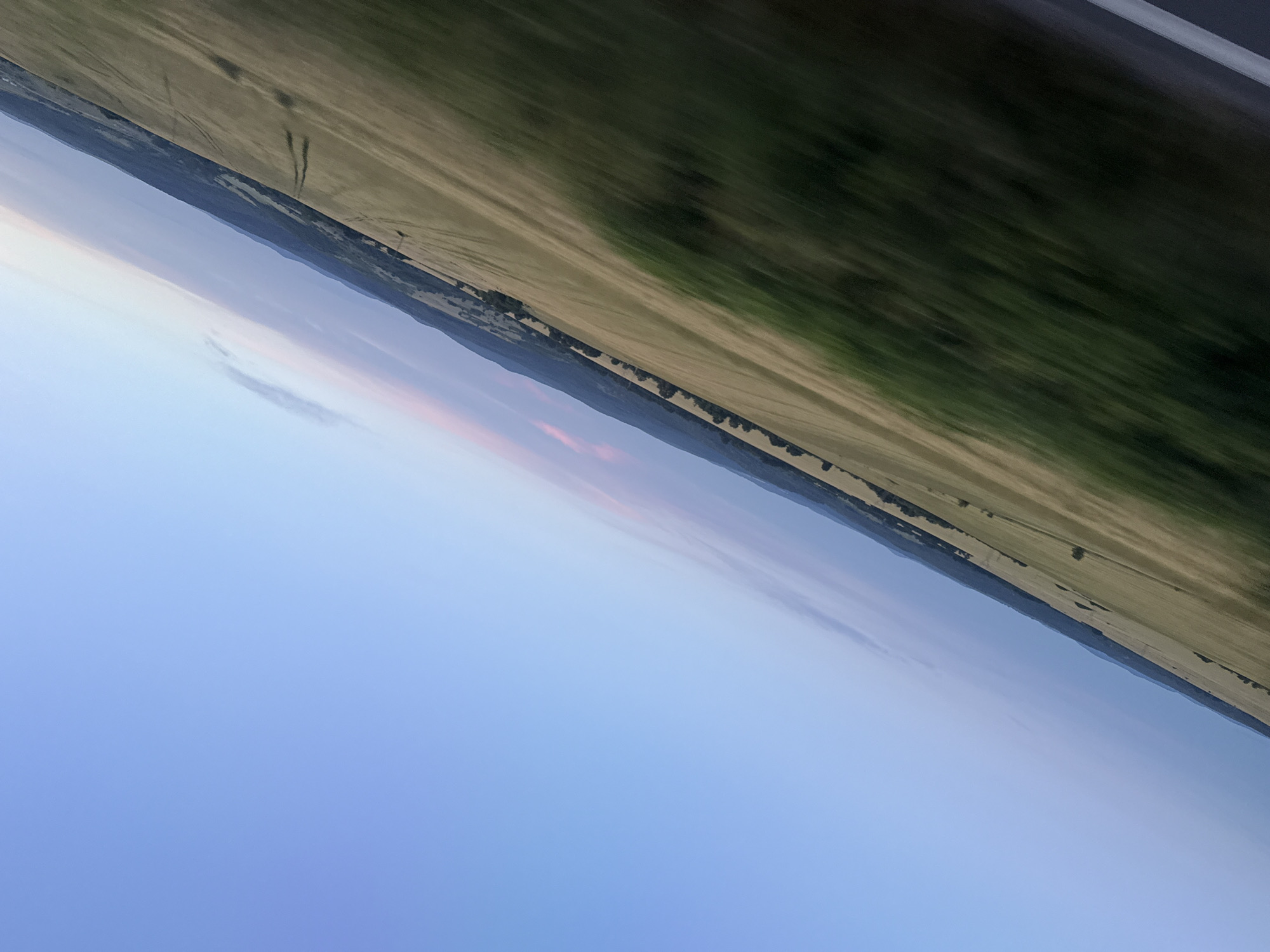 The image depicts a wide, diagonal view of a rural landscape under a clear, pale blue sky with hints of pink and purple near the horizon. The scene is tilted, suggesting it was captured from a moving vehicle. In the foreground, a blurred green field is visible, transitioning to a dry, golden field further into the distance. The golden field shows subtle patterns of tire tracks. Trees line the edge of the fields, forming a dark band against the horizon, which is marked by rolling hills and mountains. The overall impression is one of a vast, open countryside during what appears to be either sunrise or sunset. The location is specified as being in the city of Izvor, Bulgaria.