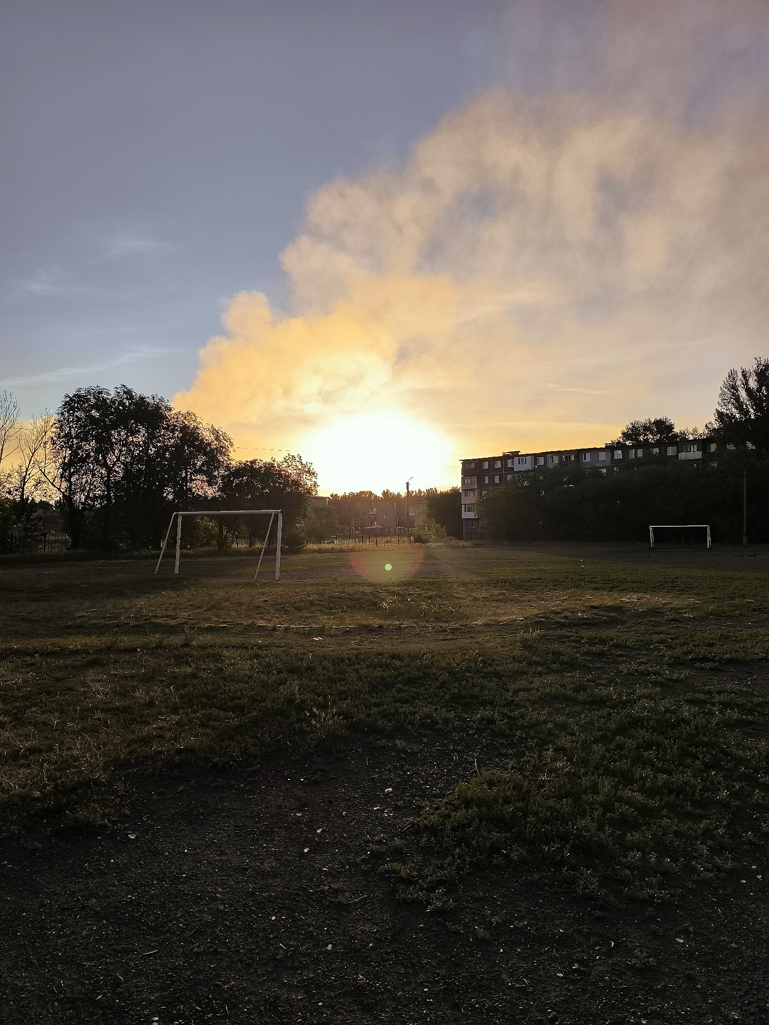 The image depicts an outdoor scene at dusk, likely in an urban area of Qarağandı city administration, Kazakhstan. A large, multi-story building with numerous windows is visible in the background, partially obscured by trees. In the foreground, a grassy field, possibly a sports field, is present, with a white soccer goal in the left mid-ground and another in the right mid-ground. The sky is illuminated by a warm, orange sunset, with clouds catching the light. A distinct lens flare is visible in the center of the image. The overall atmosphere is one of calm and the end of the day.