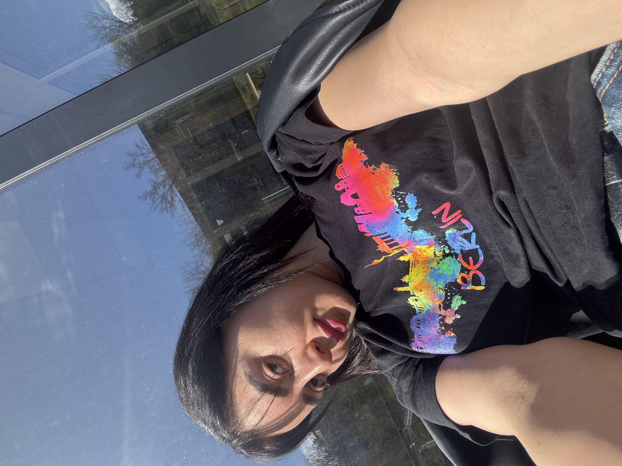 A woman is posing for a selfie. She is wearing a black t-shirt with a colorful graphic on it that says "Berlin." She also appears to be wearing a dark-colored jacket. The background shows a building with large windows reflecting the sky and trees. Based on the architecture reflected in the windows and the type of vegetation, the location is likely Tbilisi, Georgia.