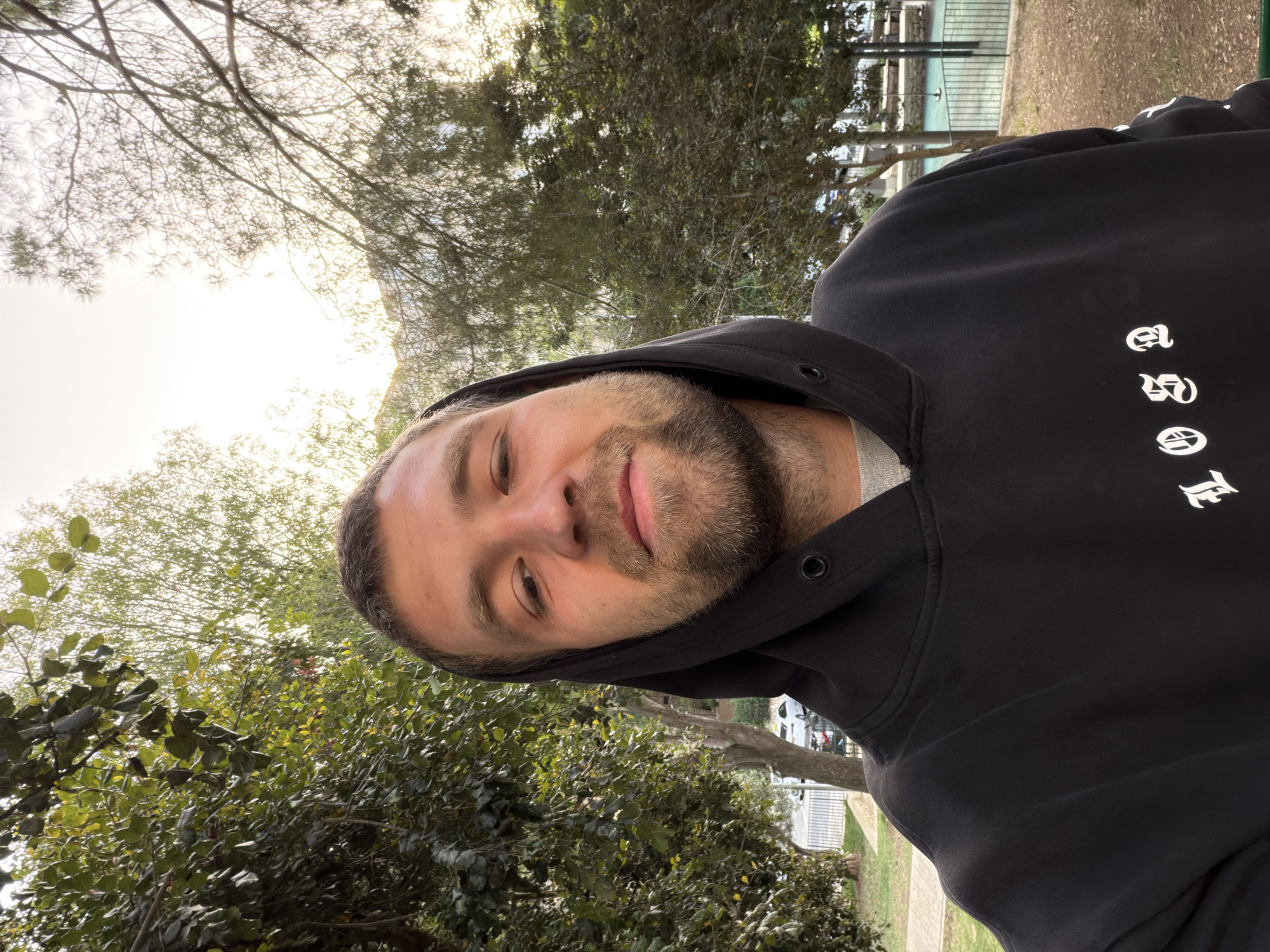 A man with a beard and wearing a black hoodie is facing the camera. He appears to be outdoors in a park-like setting with trees and a grassy area visible in the background. Part of a fence and a walkway can also be seen. The image appears to have been taken during the day, with natural light.