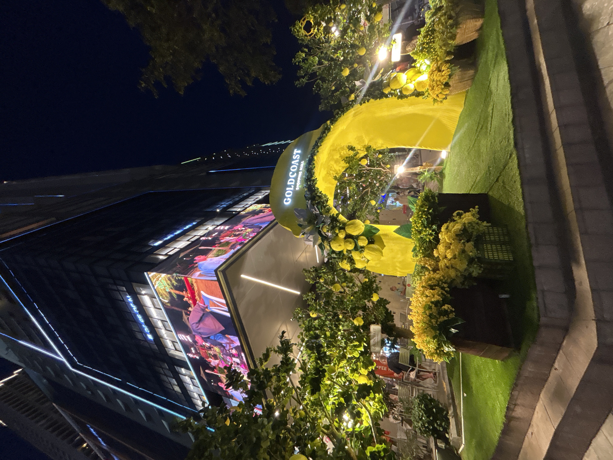 This nighttime image captures a vibrant and festive entrance to a shopping mall, illuminated by numerous artificial lights. The scene is dominated by a large, bright yellow archway that appears to be part of a decorative display, possibly themed around citrus fruits like lemons. The archway prominently displays the text "GOLDCOAST SHOPPING MALL" in white lettering.
The yellow archway is adorned with clusters of artificial lemons and green foliage, extending into trees and planters around the entrance, creating a lush and cheerful atmosphere. Several warm-toned lights are integrated into these decorations, casting a glow on the foliage and the yellow fabric of the arch.
To the left, a modern, multi-story building, presumably the Goldcoast Shopping Mall, stands against the dark night sky. Its facade features linear blue lights and a large, brightly lit digital screen displaying colorful imagery, possibly scenes with people or promotional content. On the ground level, below the archway and behind some of the decorative trees, outdoor seating with tables and chairs is visible, suggesting a cafe or restaurant area. A few indistinct figures of people can be seen in the background, suggesting activity in this area.
To the right, a patch of artificial green grass lines a paved walkway or steps, contributing to the landscaped appearance of the entrance. Planters filled with yellow flowers and more green foliage are also present. The overall impression is one of a welcoming and lively commercial space after dark, likely in Nha Trang, Vietnam, as indicated by the location context.
