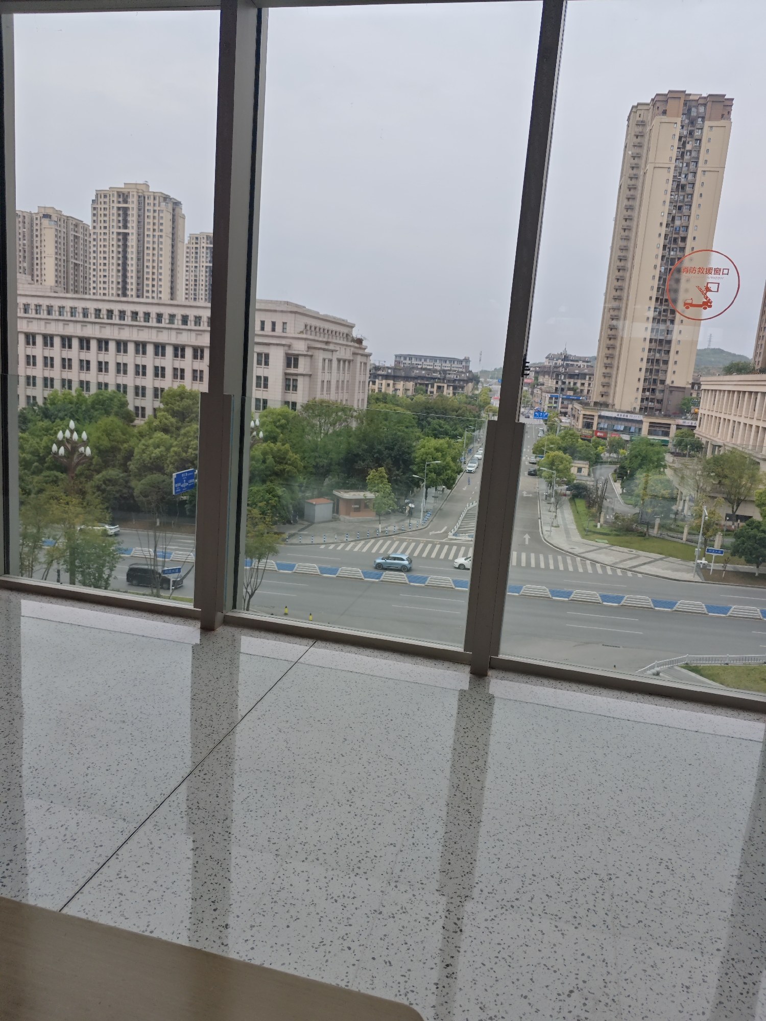 A view from inside a building looking out onto a cityscape in Liangjiang, China. The foreground shows a light-colored terrazzo floor that reflects the outside view. Through the window, there is a street with multiple lanes and pedestrian crossings. Various buildings are visible, including high-rise apartments and a large, multi-story building with classical architectural elements. Green trees line the streets and fill the spaces between buildings. The sky is overcast. Traffic is visible on the roads. A red and white circular sticker is on the window in the upper right area of the photo.