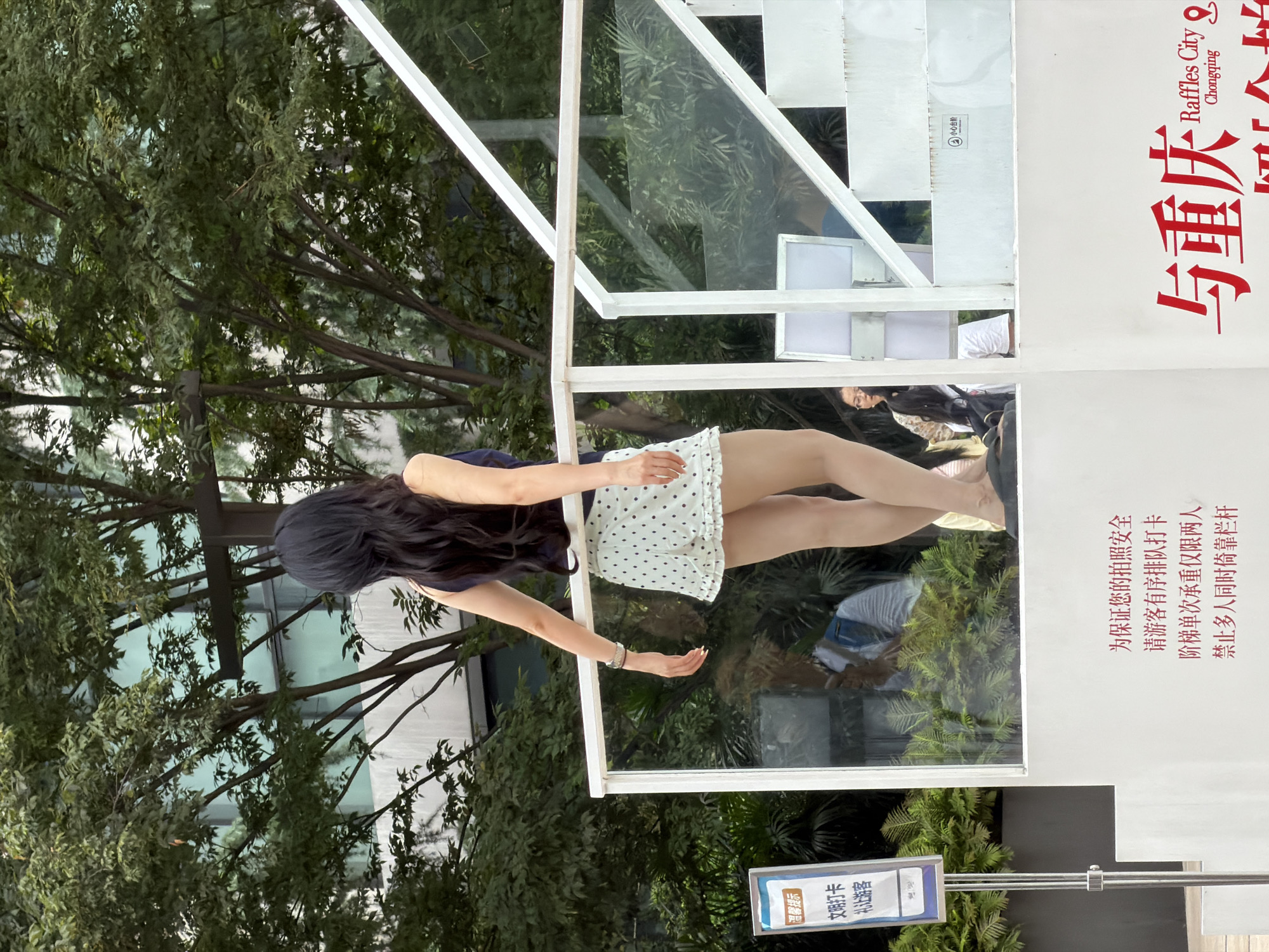 In an outdoor urban setting, likely Chongqing, Yuzhong District, China, a woman with long, dark hair stands on a white platform with a glass railing. She is facing away from the camera, wearing a dark sleeveless top and a white skirt with polka dots. Her left arm is resting on the railing, and she is wearing a bracelet on her left wrist. A sign with Chinese text is visible to her left, and reflections in the glass railing show other people and greenery. A large red text graphic that appears to say "与重庆 Raffles City Chongqing" is visible in the lower right corner. The background consists of lush green trees and the facade of a building.