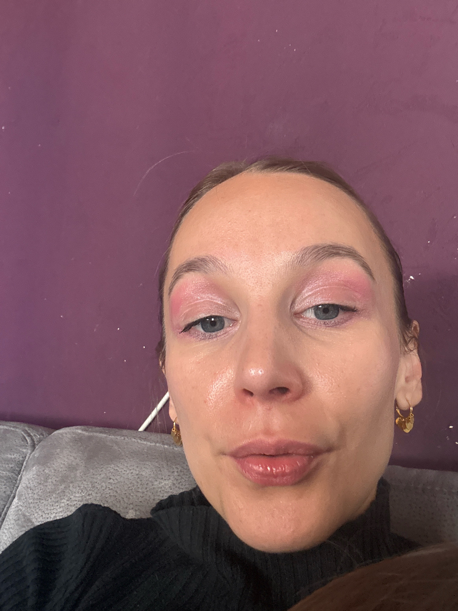 The image shows a close-up, selfie-style shot of a woman's face. She has light skin, blue eyes, and is wearing pink eyeshadow. Her hair is pulled back. She is wearing a black turtleneck. She has small, gold hoop earrings with charms. The background is a textured purple wall. A portion of a grey sofa is visible in the lower left of the frame. The image does not provide any specific information about the location within Überherrn, Germany.