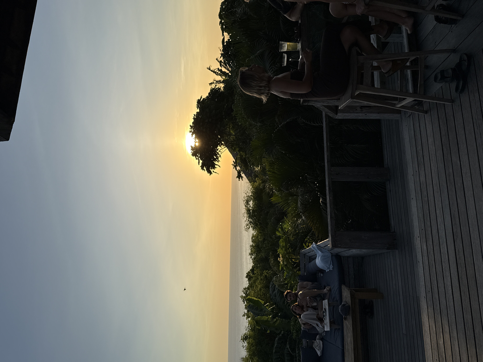 The image depicts a sunset view in Kuta Selatan, Indonesia. The sun is partially obscured by a tree, casting a golden glow over the ocean and sky. A wooden deck is visible, with people seated on chairs and a lounge area. Some individuals are sitting at a table with drinks. The surrounding environment includes lush vegetation.