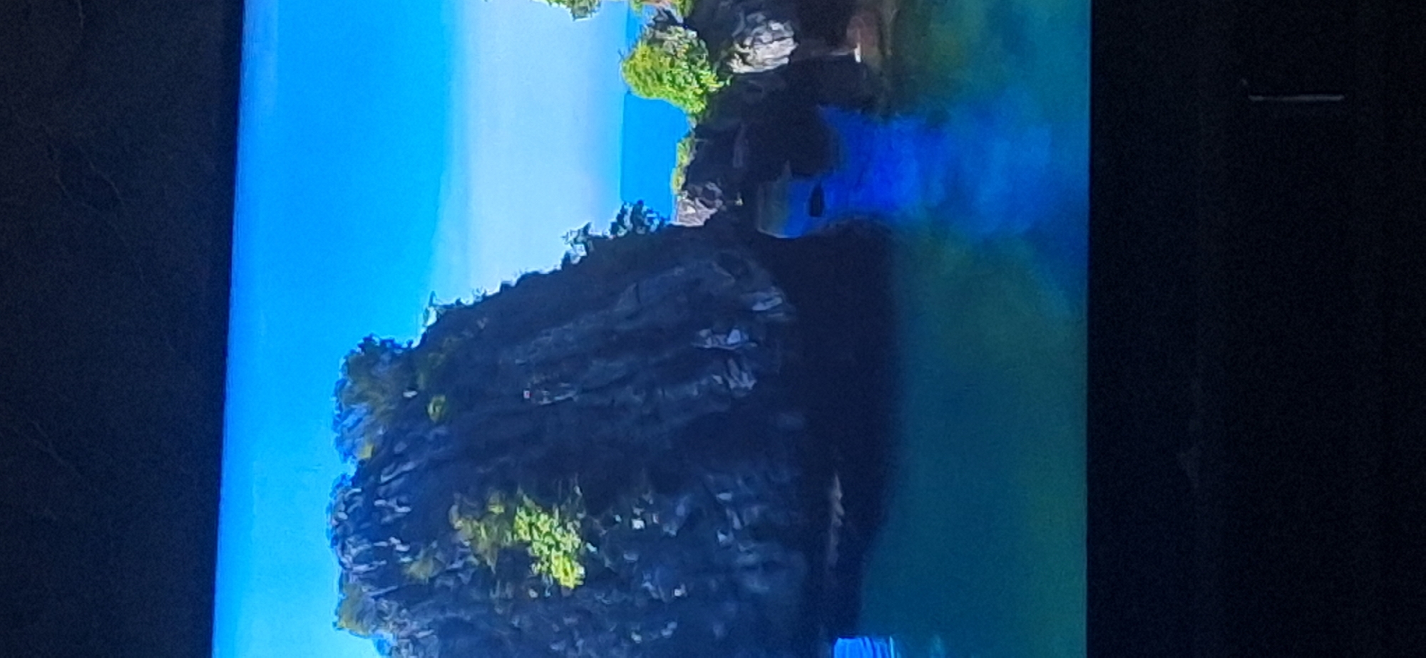 The image shows a vibrant blue sky and clear water surrounding rocky formations covered with lush green vegetation. The water reflects the blue sky and the dark rocks, creating a striking contrast. The scene appears to be a natural landscape, possibly a coastal area or a body of water with islands. There is no visible evidence of the city of Bila Tserkva, Ukraine, or any human presence. The image is a still photograph.