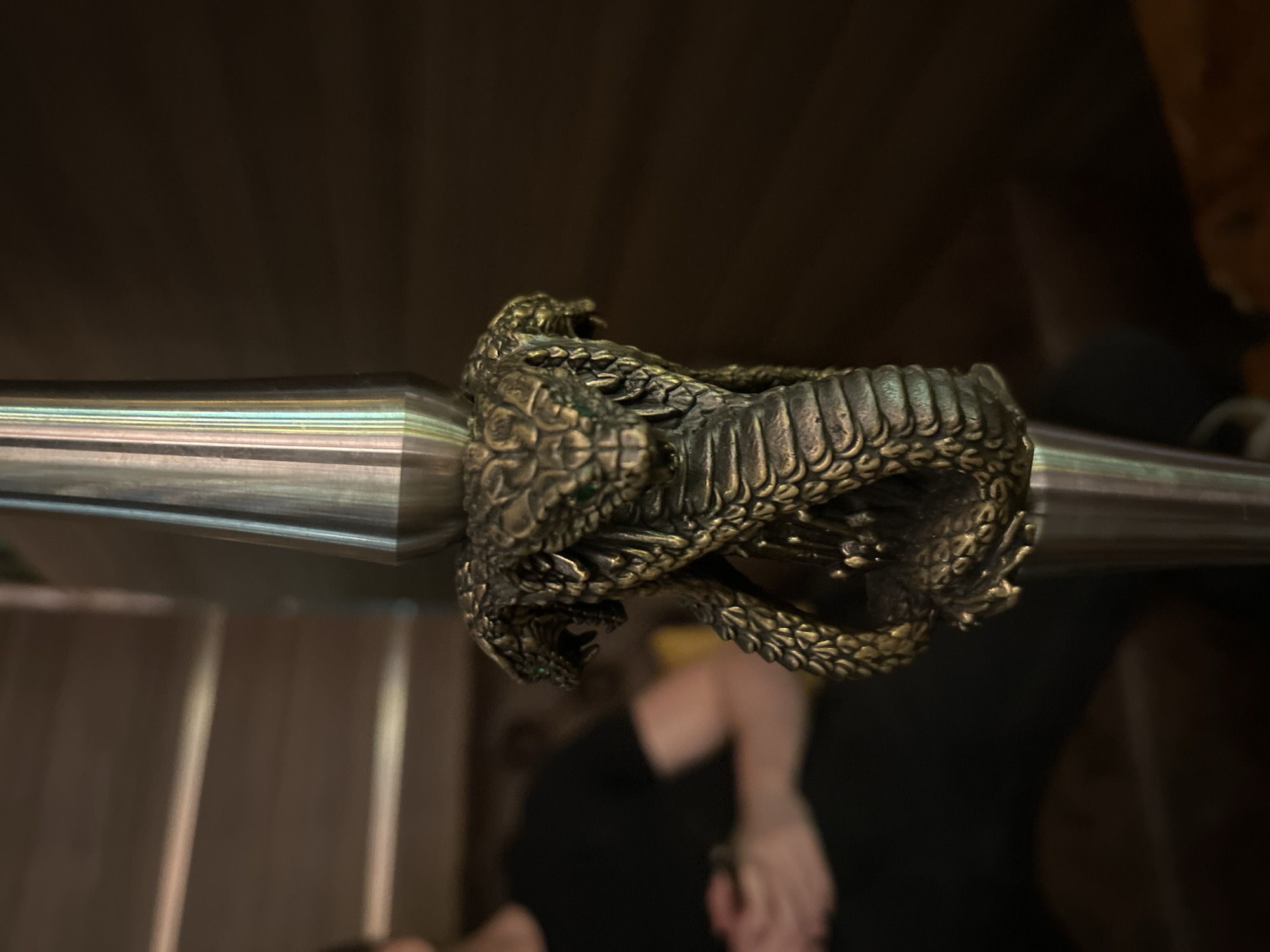 A close-up, medium shot captures a decorative hookah stem featuring an intricately carved brass cobra entwined around its base. The cobra's scales, fangs, and eyes are detailed, with small green accents in its eye sockets. The stem itself is made of brushed metal and has a cylindrical shape. In the blurred background to the left, a person wearing a black t-shirt and dark pants is seated, with their hands clasped in their lap. The environment appears to be indoors, possibly a lounge or a room with wooden paneling on the walls and stairs. The lighting is dim and warm, casting shadows and highlighting the metallic sheen of the hookah stem. The scene is static and focused on the ornate hookah detail. The specific location in Burgas, Bulgaria, is not discernible from the visual information provided.