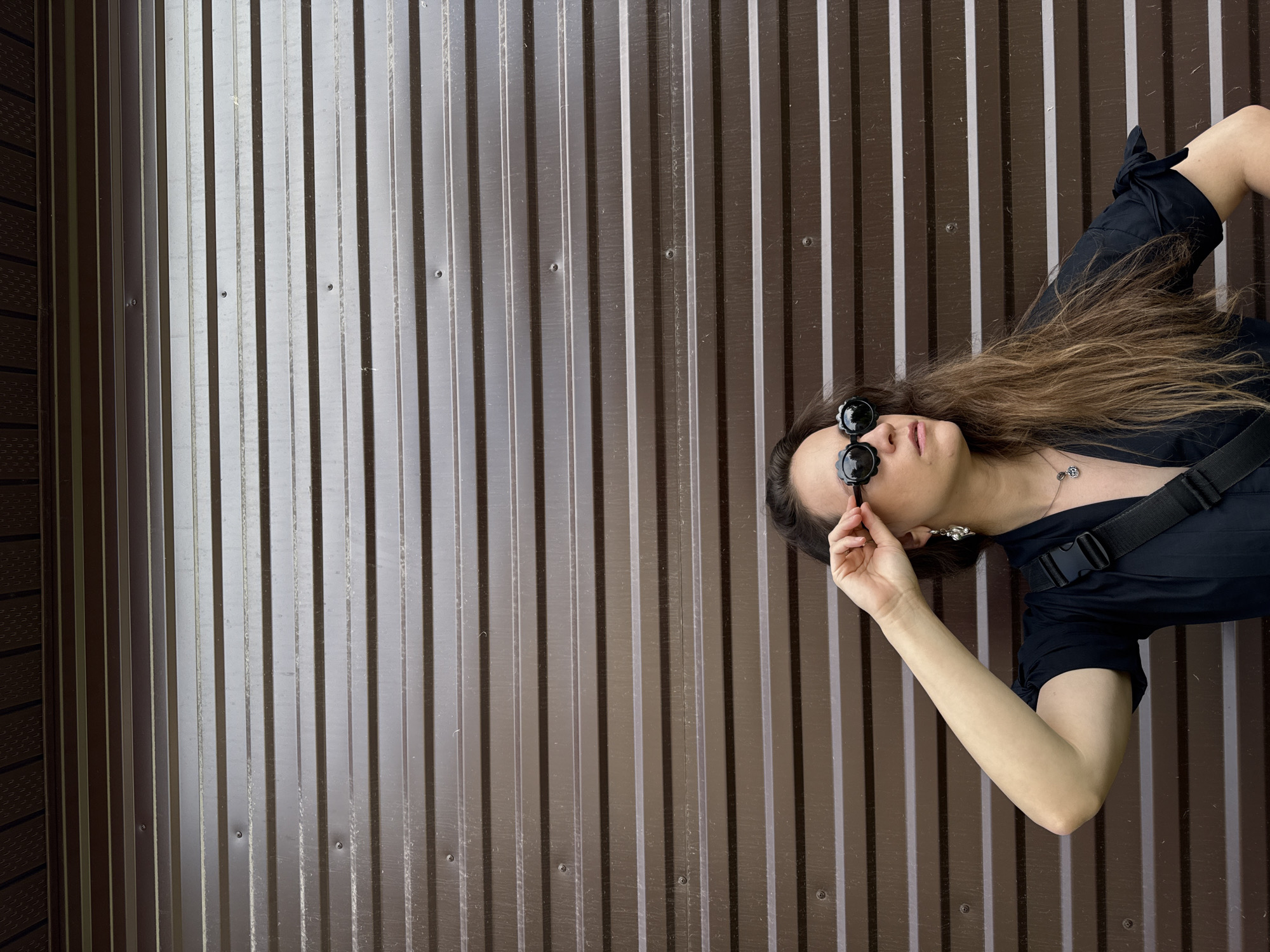 A young woman with long, brown hair lies on her back against a dark brown corrugated metal wall. She is wearing a black top, black sunglasses with round, scalloped frames, and a silver pendant necklace. Her left arm is bent at the elbow, and her hand is reaching up to adjust her sunglasses. The wall behind her has vertical ridges and appears to be outdoors. The lighting suggests it is daytime, with some sunlight casting shadows. The overall mood seems relaxed or contemplative. There is no visible text in the image. The scene is set in Chaikovsky, Russia.