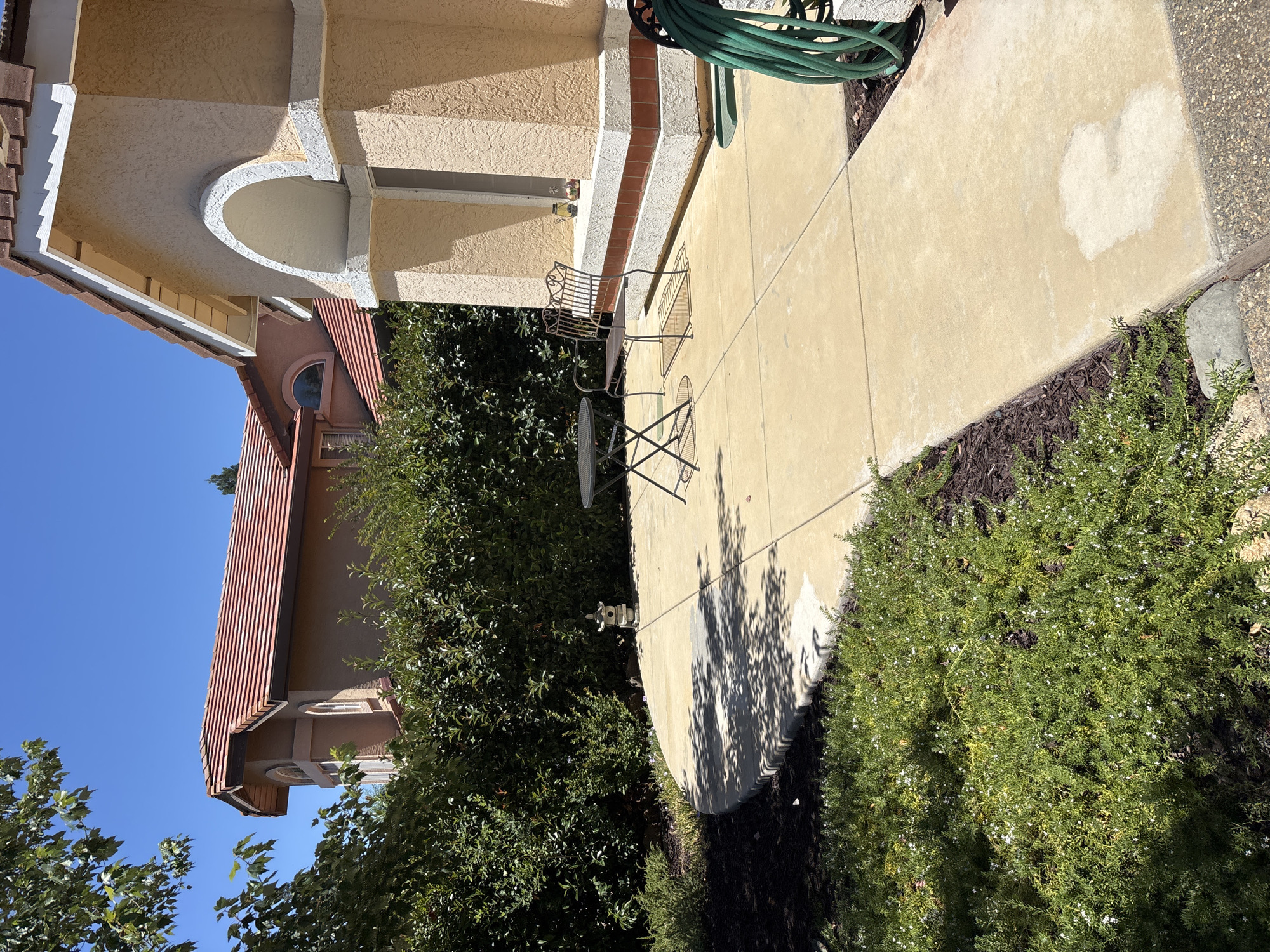 A sunny day in Folsom, United States of America, reveals a residential patio area. The patio is paved with light-colored concrete tiles and bordered by dark mulch and lush green bushes. A small, dark metal bistro set, consisting of a round table and two chairs, sits on the patio, casting dappled shadows. To the right, a dark metal bench with intricate scrollwork is placed near the house. The house features a light yellow stucco exterior with a white arched entryway and a red tile roof. Adjacent to the house, a coiled green garden hose is visible. The scene is set against a backdrop of dense green foliage and a clear blue sky.