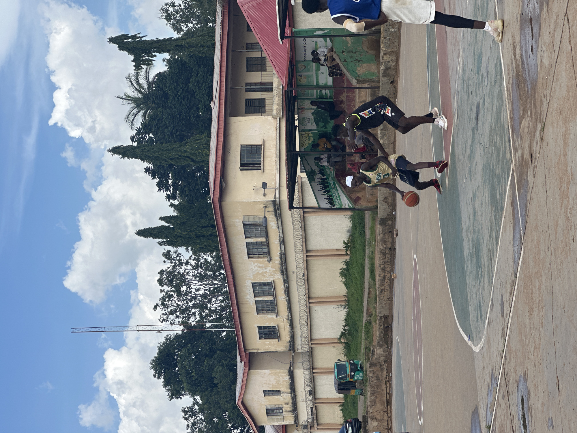 The image depicts an outdoor basketball court in Jos, Nigeria. In the foreground, three young men are engaged in a basketball game. One player, wearing a yellow jersey with the number "3" and dark shorts, is dribbling the ball. Another player, in a black jersey with colorful accents and black shorts, is defending. A third player, partially visible on the right, is wearing a blue jersey and white shorts. The court itself appears to be made of concrete with painted lines marking the boundaries and key areas. In the background, a light-colored, two-story building with a red roof stands behind a beige-colored wall topped with barbed wire. Adjacent to the building is a set of green bleachers with spectators seated. A small, green and yellow auto-rickshaw is parked to the left of the building. Trees and a tall antenna tower are visible against a cloudy sky. The overall scene suggests a casual basketball game taking place in a community or recreational area.