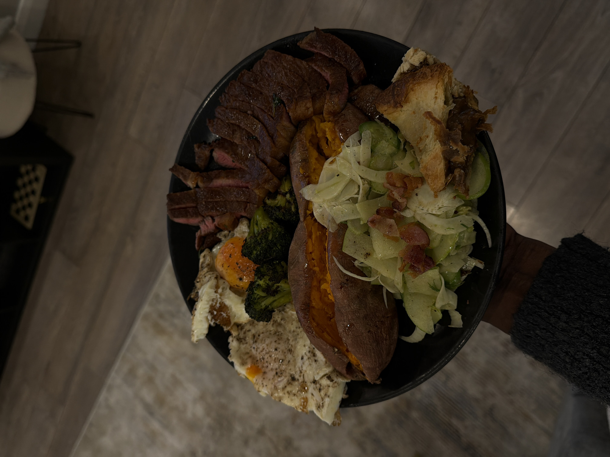 A person is holding a black plate with various foods. The plate contains sliced steak, a halved sweet potato, fried eggs, broccoli, and a salad consisting of greens and bacon. The plate is being held in front of a blurred-out background, which appears to be a room with light-colored flooring. The person holding the plate has dark skin and is wearing a gray sweater. The image does not provide enough information to determine if it was taken in London or to identify any specific location.
