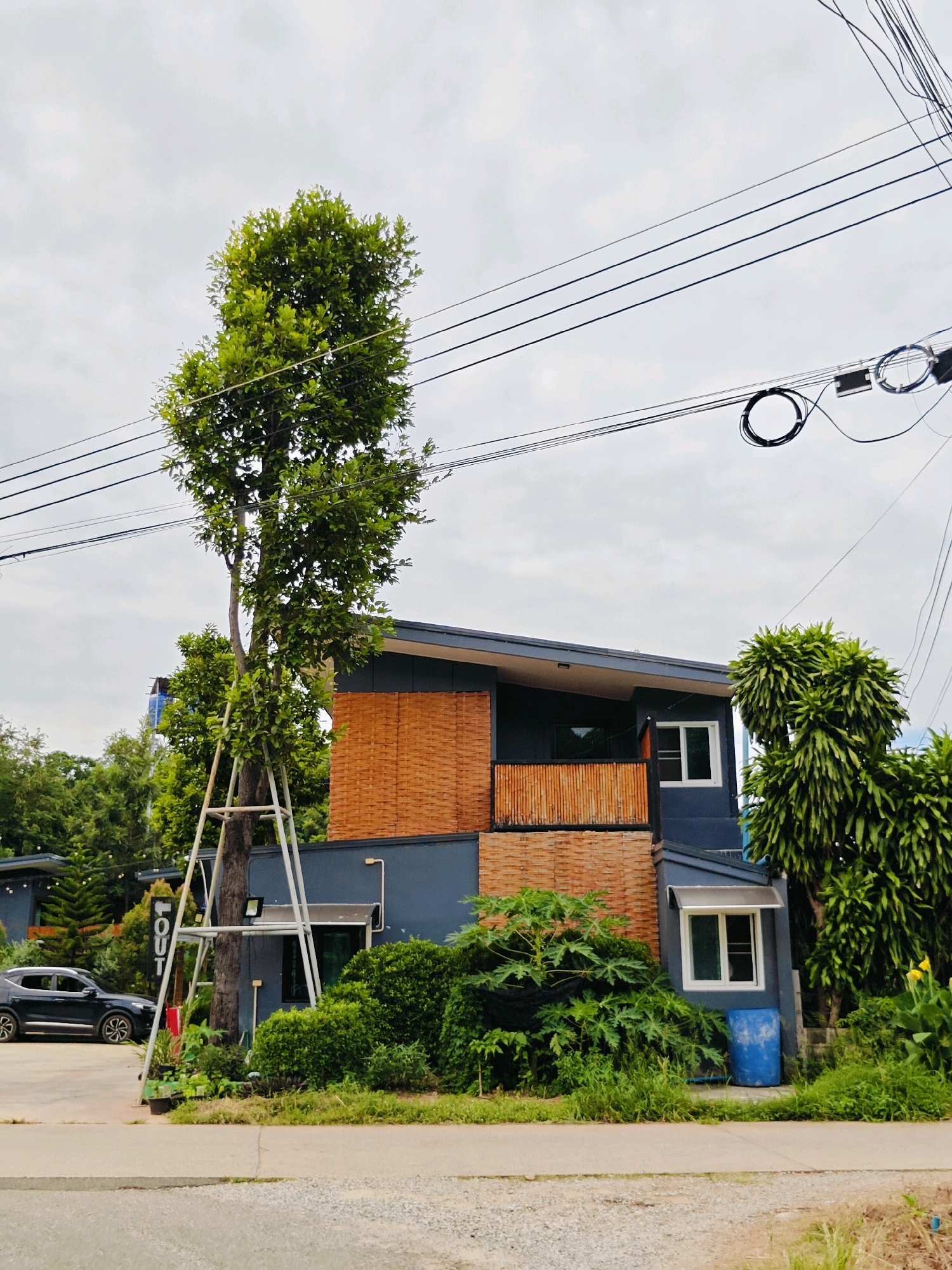 The image depicts a street scene in Mu Si, Thailand, on an overcast day. The focal point is a two-story modern building with dark blue walls and sections of woven bamboo-like material. A tall, leafy tree, supported by a metal frame, stands prominently in front of the building. The ground level features lush green foliage and a blue barrel. To the left, a dark-colored SUV is parked on a paved area. Overhead, numerous power lines are strung across the sky. The environment appears to be a mix of residential and potentially commercial, with dense greenery surrounding the structures.