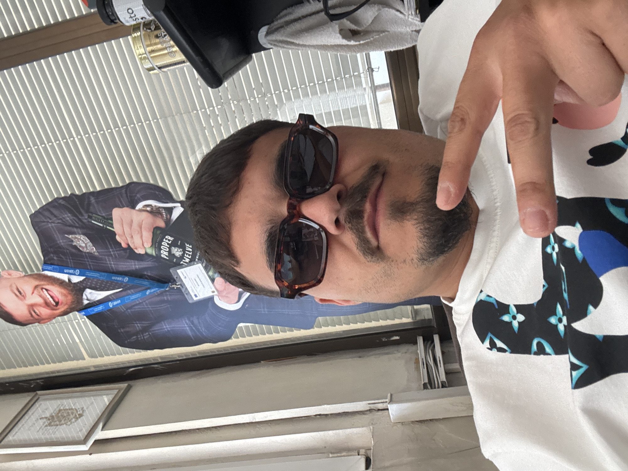 A man with dark hair, a mustache, and sunglasses is posing for a selfie. He is wearing a white t-shirt with a black and blue design. He is holding up two fingers in a "peace" sign. In the background, there is a cardboard cutout of Conor McGregor holding a bottle of Proper Twelve whiskey. The photo was likely taken in Plovdiv, Bulgaria, as evidenced by the "Rason" lanyard on the cutout of McGregor, which is a Bulgarian company based in Plovdiv. Other details include the presence of a window with blinds, a framed picture on the wall, and what appears to be a container labeled "Pesto" near the top of the frame.
