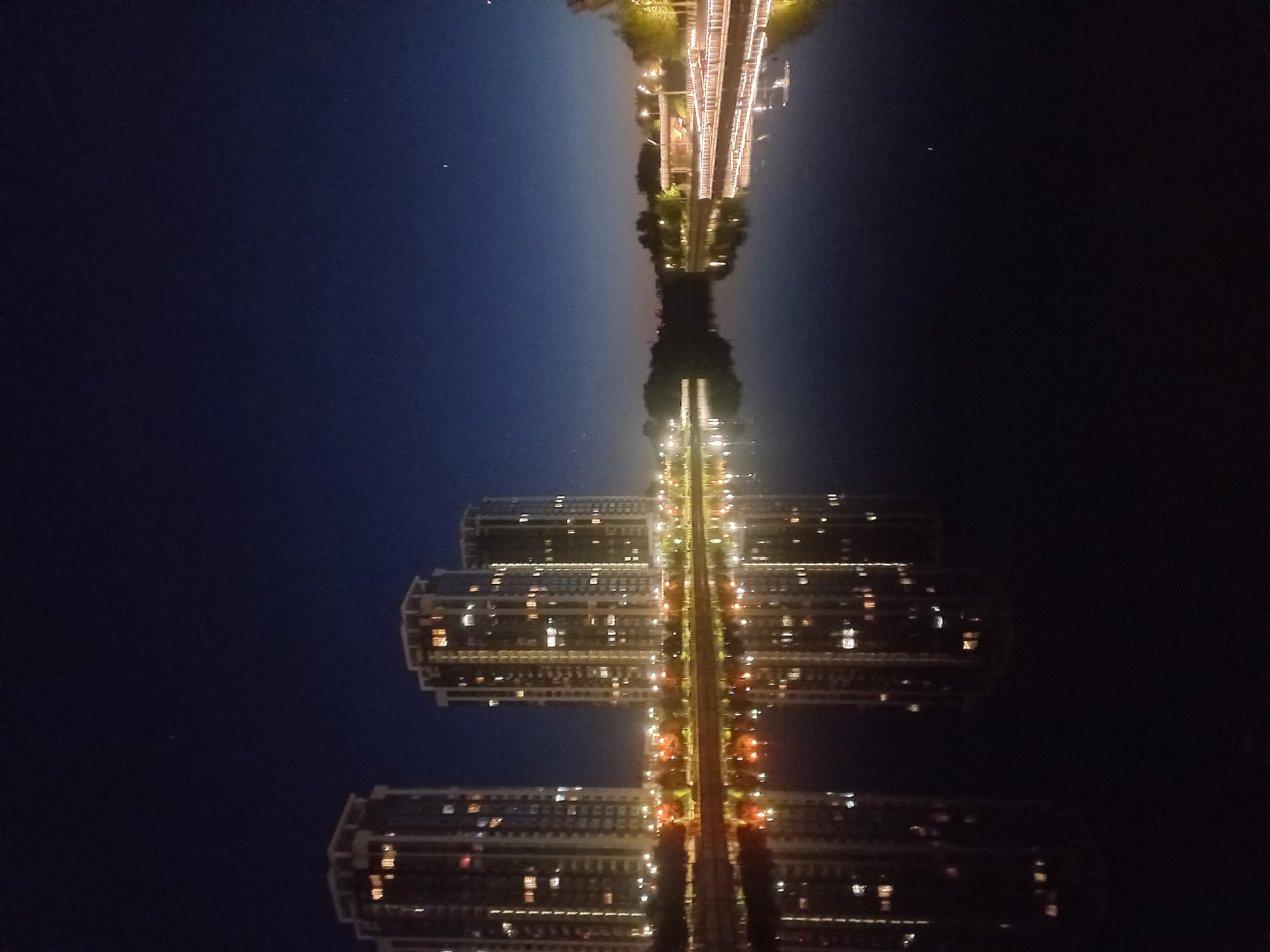 The image shows a reflection of a nighttime scene in water. The reflection includes illuminated buildings resembling residential complexes and a central illuminated walkway. The upper portion of the image, also reflected, appears to show a park or green area with a brightly lit elevated structure. The sky is dark blue, with some faint stars visible. Based on visual indicators, the location is likely Kyiv, Ukraine, particularly the area of the Obolon district near the Dnieper river.