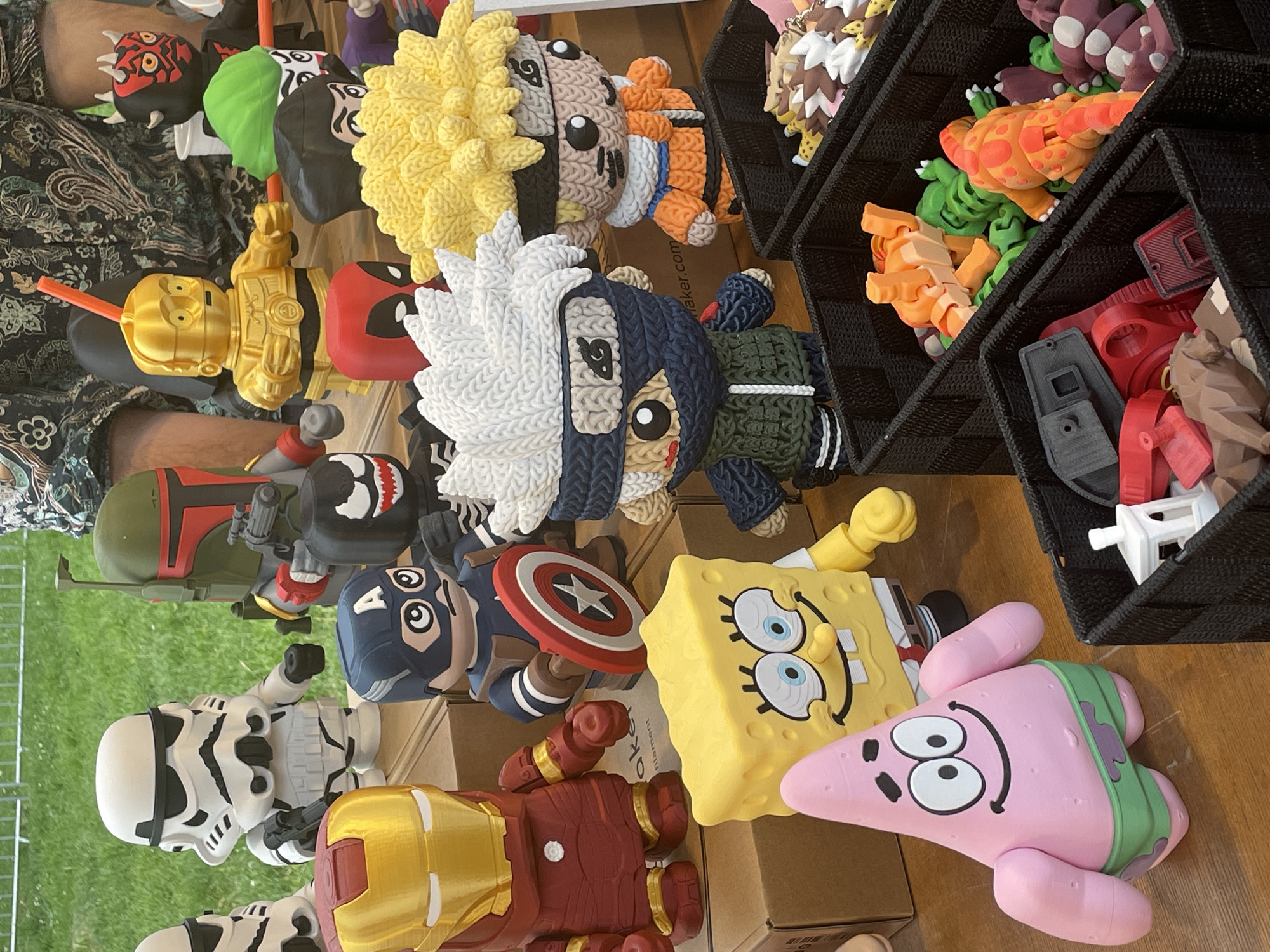 The image depicts a display of various figurines and toys, possibly at a market or fair. Notable characters represented include Stormtroopers, Iron Man, Captain America, Boba Fett, Spongebob Squarepants, Patrick Star, C-3PO, Deadpool, Naruto, and Kakashi. Some items are arranged on cardboard boxes, while others are in black woven baskets containing articulated animals and other objects. The location is likely in Zemun Urban Municipality, Serbia, based on prior knowledge. The scene suggests a vendor selling pop culture-themed merchandise.