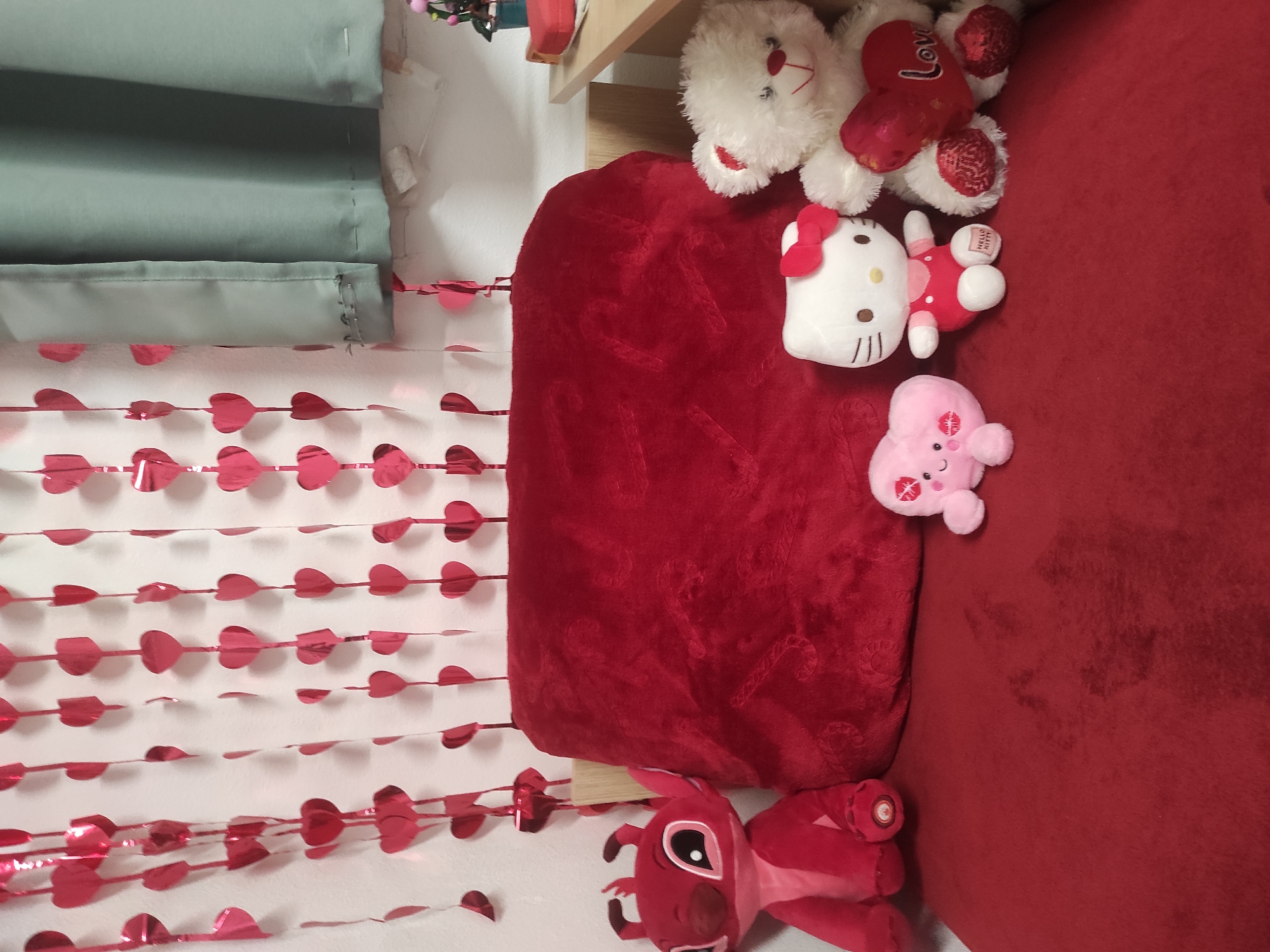 The scene depicts an interior setting adorned with Valentine's Day decorations. A string of red, heart-shaped foil garlands hangs vertically against a white wall, partially obscured by a pale green curtain. A wooden shelf is visible, holding an assortment of plush toys on a red, heart-shaped cushion. These include a white teddy bear, a red "LOVE" bear, a Hello Kitty plush, and a pink, heart-shaped plush with a face. Below the cushion, another red plush toy is partially visible. The overall environment suggests a celebratory or romantic theme. There is no indication of the location in Sofia, Bulgaria or any people.