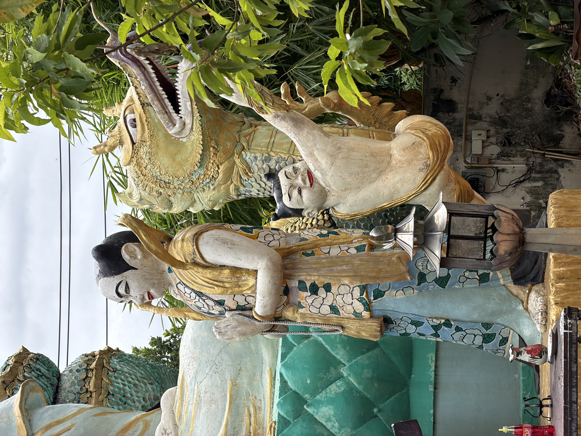 This image depicts a religious shrine or temple in Chang Phueak, Thailand. The scene features several statues, including a large, serene figure dressed in traditional garments, possibly a deity or a revered person, with hands clasped in prayer. To the right of this figure is a dynamic sculpture of a dragon, its head raised and mouth open, adorned with golden scales. In front of the dragon, a smaller statue of a playful child-like figure is depicted in a dynamic pose, reaching upwards. The statues are richly decorated with gold and vibrant colors, suggesting significant artistic craftsmanship.
In the foreground, a small table holds a red bottle of soda, a decorative vase with floral motifs, and a small dish. A metallic lantern with glass panes is placed on a pedestal to the right of the table. The background is dominated by lush green foliage and a cloudy sky. The overall atmosphere suggests a place of spiritual significance and cultural richness within the urban landscape of Chang Phueak.