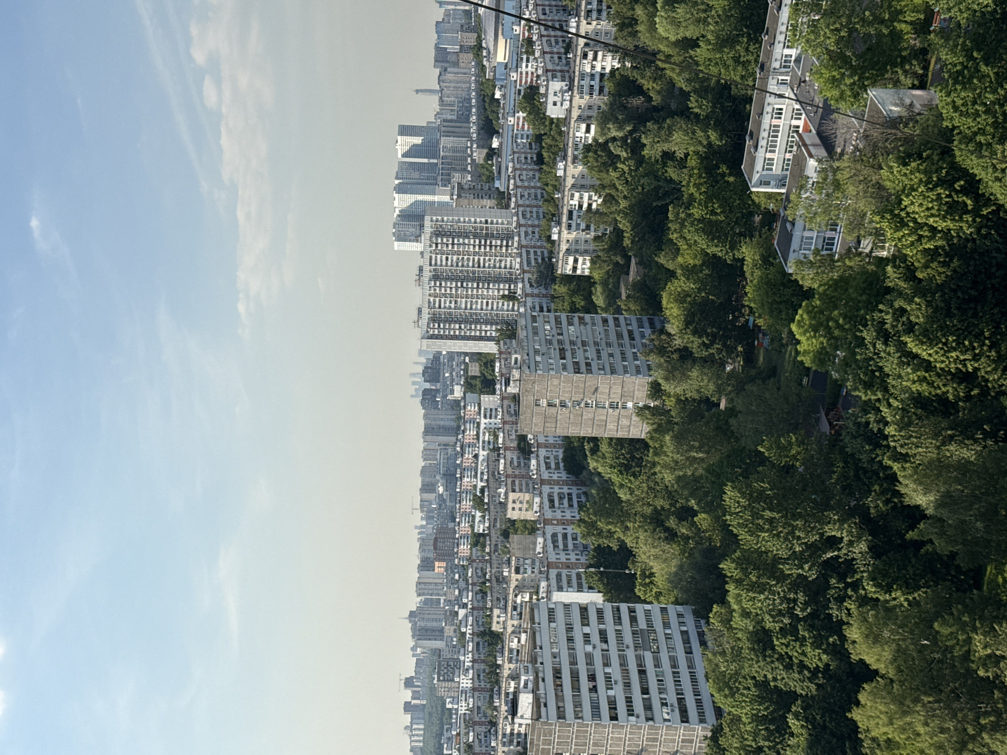 This image presents a high-angle, panoramic view of a densely populated residential area within Moscow, Russia, captured under a hazy light blue sky with sparse, wispy clouds. The scene is dominated by a mix of Soviet-era architecture and abundant mature greenery, indicating a well-established urban landscape.
In the foreground and midground, numerous multi-story apartment buildings are visible. To the immediate left, a large, grey, rectangular panel building with many uniformly arranged windows stands prominently. Further into the midground, slightly to the right of the center, another tall, slender, grey panel building is situated amidst the trees. The area is heavily wooded, with dense clusters of deciduous trees in full leaf, suggesting parks, courtyards, or tree-lined streets throughout the residential blocks. Scattered smaller, typically 5-9 story, light-colored or pastel-toned apartment buildings are visible beyond the immediate foreground.
A distinctive landmark in the middle distance, slightly left of the image's center, is a large, modern, multi-sectional white residential complex, identifiable as likely the **Aerobus Residential Complex**. Beyond this, the cityscape extends towards the horizon, where the distinct and prominent silhouette of the **Moscow International Business Center (Moscow City)**, with its cluster of modern skyscrapers, is clearly visible in the far distance, slightly to the right of the central axis.
No individuals or specific actions are discernible due to the elevated perspective. A black diagonal cable or wire runs from the top right corner down towards the lower right.
Based on the visible landmarks – specifically the Aerobus Residential Complex in the mid-distance and the Moscow City skyline in the background – this photograph was taken from an elevated vantage point in a residential district located **north-east of the Aeroport District** of Moscow, likely within the **Koptevo, Timiryazevsky, or Voikovsky Districts**, looking towards the south-west.
