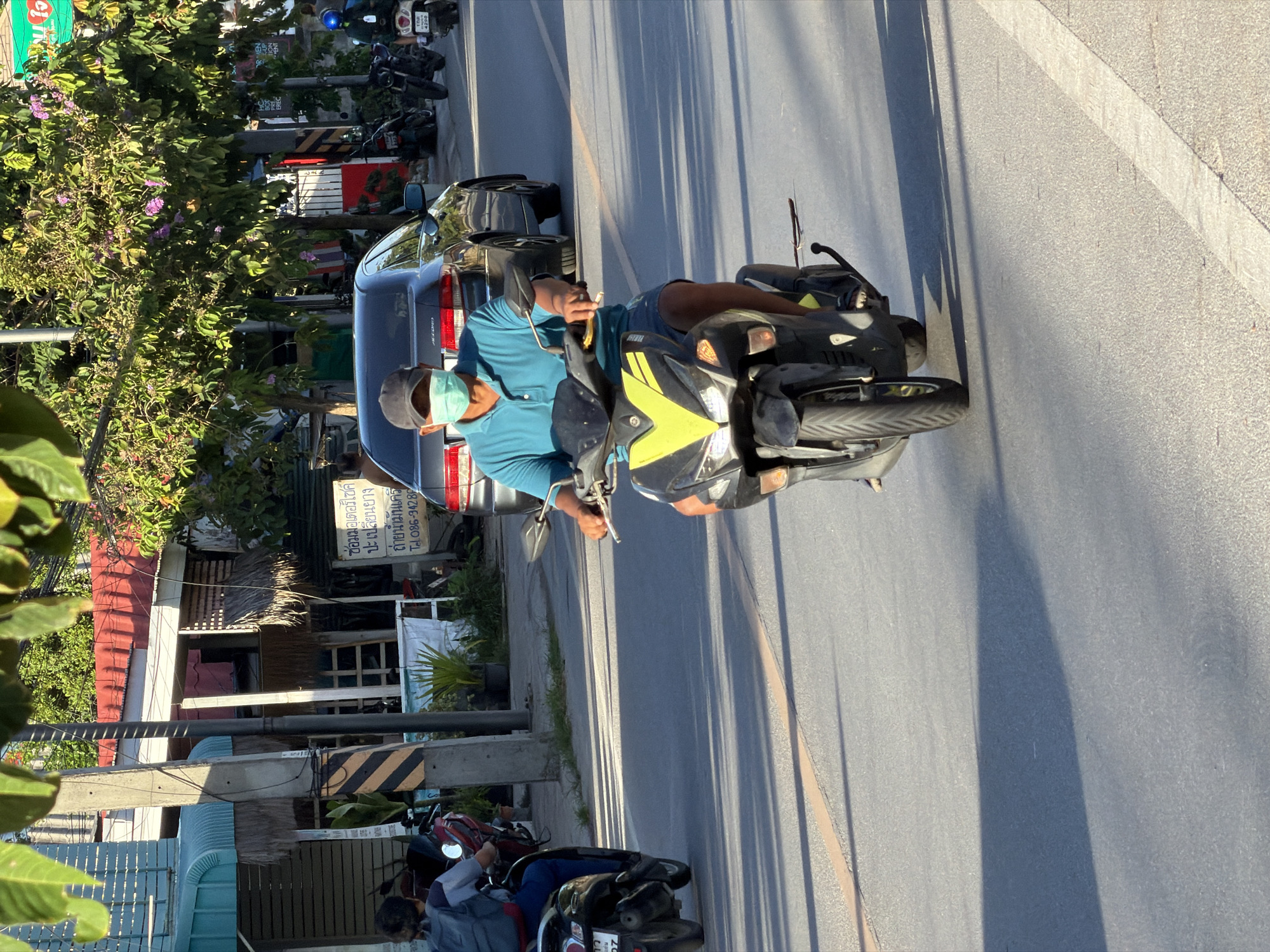 A man wearing a blue polo shirt, blue shorts, a black baseball cap, and a light blue face mask rides a black and yellow scooter on a paved road in Ko Samui District, Thailand. The scooter has a license plate that reads "4200". The man is holding the handlebars with his right hand and appears to be looking forward. The road is lined with businesses and trees. In the background, a dark gray sedan is parked on the side of the road. Other motorcycles are also visible parked nearby. The sun is casting long shadows on the road, suggesting it is late afternoon. The scene is typical of a street in a Thai town, with a mix of vehicles, pedestrians, and commercial establishments.