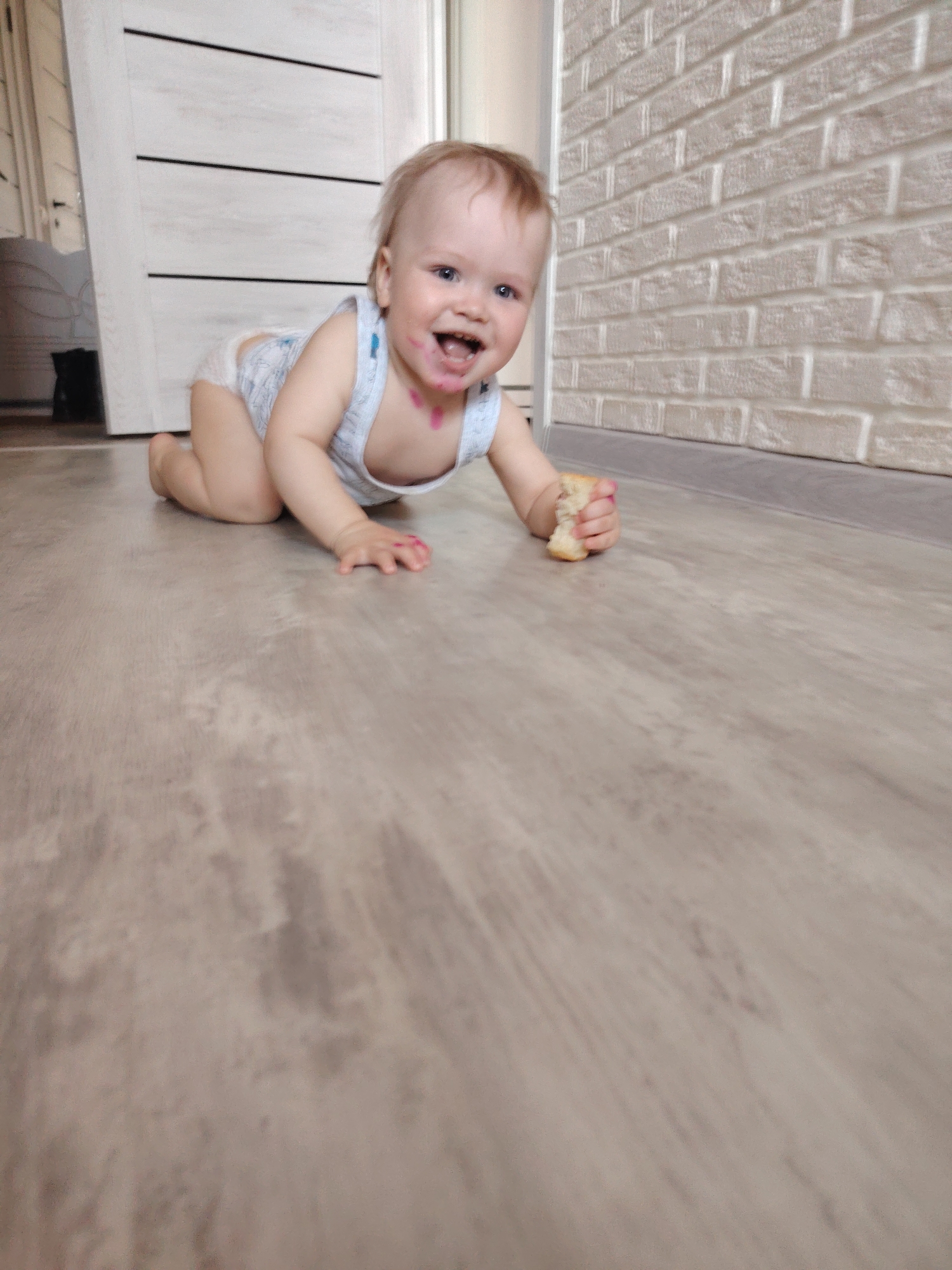 A toddler is crawling on a beige floor in a room in Chaikovsky, Russia. The toddler has some food smudged around its mouth, and is smiling while holding a piece of food in its hand. The room has a brick wall on one side and a white door on the other side.