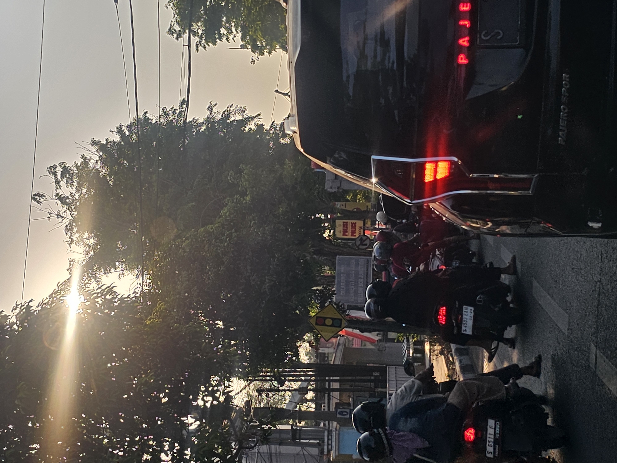 The image depicts a street scene in Tuban, Indonesia, during sunset or sunrise. The sun's rays are visible, casting a warm glow and lens flare through the leaves of trees lining the road. Several motorcycles, occupied by riders wearing helmets, are seen in the foreground and middle ground, suggesting traffic. One motorcycle in the foreground has a license plate reading "S 6511 F". Another motorcycle behind it has a license plate reading "S 4503 EAH". In the far right of the image, the rear of a dark-colored SUV is prominently displayed, showing its red taillights and the word "PAJERO" illuminated in red on the tailgate. Below that, text reads "PAJERO SPOR". A traffic light signal is visible on a pole to the left, showing yellow and red lights. There are also signs, including one with the word "POLISI" repeated, and another that appears to be a "YELLOW BOX JUNCTION" sign. The environment suggests an urban setting with buildings and power lines visible in the background.