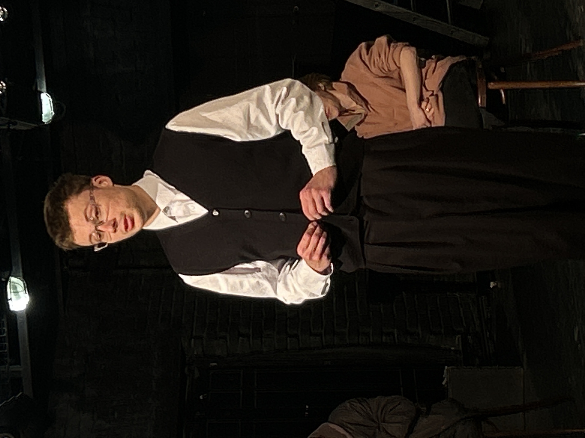 An indoor, vertically oriented image features two individuals in what appears to be a theatrical setting. The primary subject is a young man with short brown hair and glasses, wearing a white collared shirt, a dark vest, and dark trousers. He stands in the foreground, facing slightly left, with his hands positioned near his waist, seemingly adjusting the buttons of his vest. His mouth is slightly agape, and his expression is focused or intense.
Behind him and to the right, a second person is partially visible, slumped over what appears to be a wooden chair. This individual wears a light brown or peach-colored long-sleeved shirt and dark pants, with their head resting down and away from the camera, suggesting they are asleep, unconscious, or in a state of exhaustion.
The background is dark and indistinct, featuring dark, possibly brick-patterned walls and a floor. Stage lighting fixtures with a soft glow are visible on the left side of the frame, illuminating parts of the scene. The overall ambiance is dim and dramatic, suggesting a moment from a play, rehearsal, or performance. No visible text is present in the image.