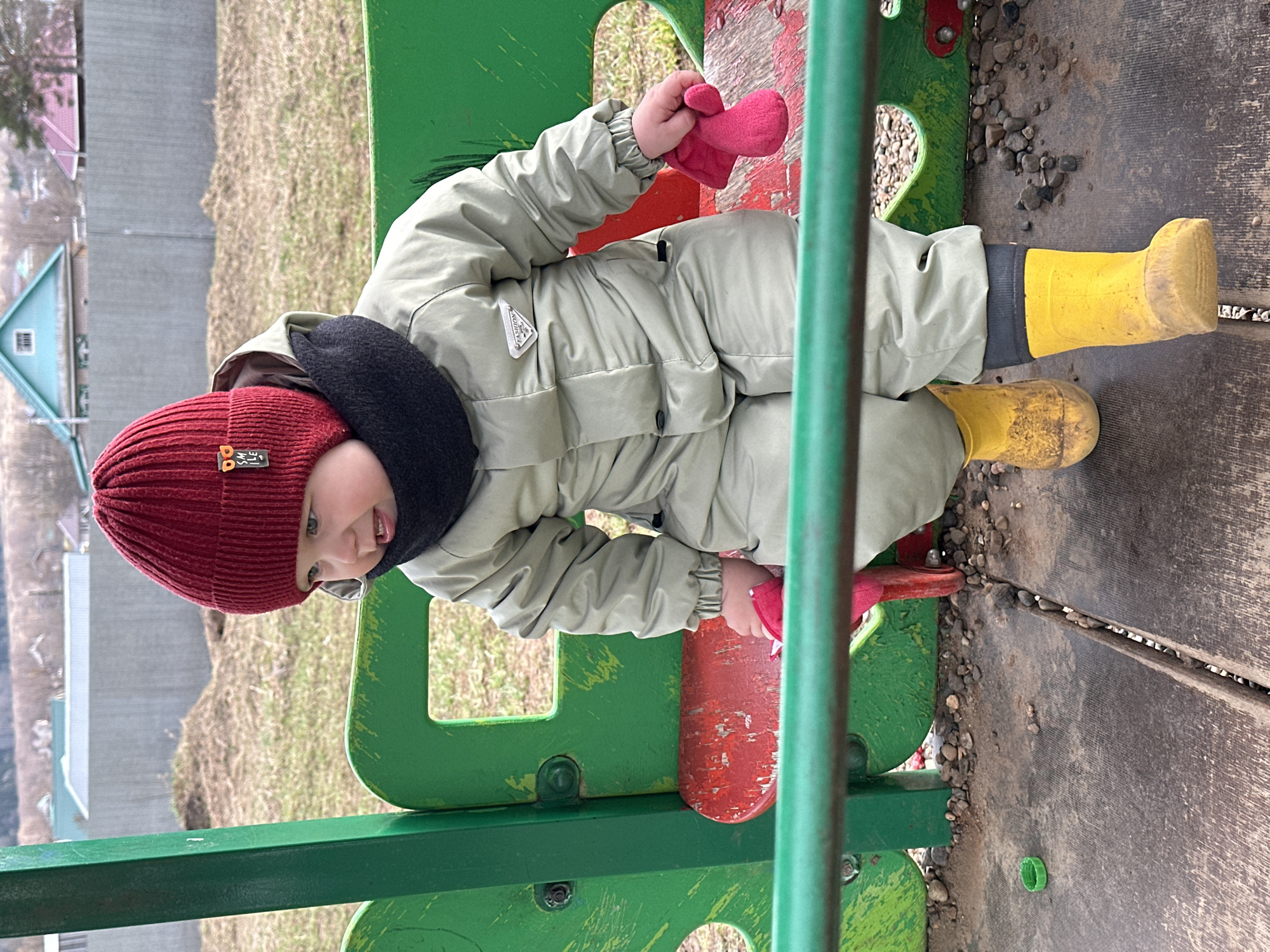 A small child, wearing a maroon knit cap, a black neck warmer, a light grey snowsuit, pink mittens, and yellow rain boots, sits on a green and red seesaw. The seesaw is on a wooden surface with gravel nearby. In the background, there's a grassy area and a building with a blue roof. The child is smiling and looking towards the viewer. The location is Ludorvay, Russia.