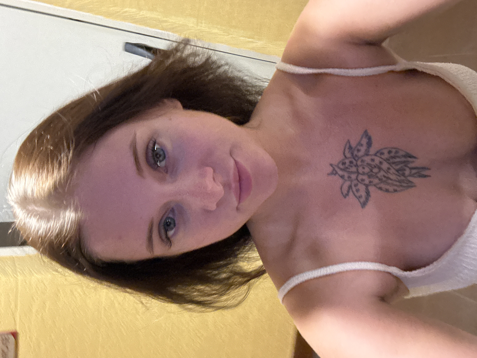 A selfie taken from a chest-up perspective shows a young woman with light brown hair, which is partially illuminated and appears lighter on top. She has blue eyes and fair skin. A tattoo of a stylized flower is visible on her chest, below her neck and above her cleavage, which is partially exposed by her white, ribbed tank top. The background consists of a yellow, textured wall on the left and a white door on the right. The scene is indoors.
