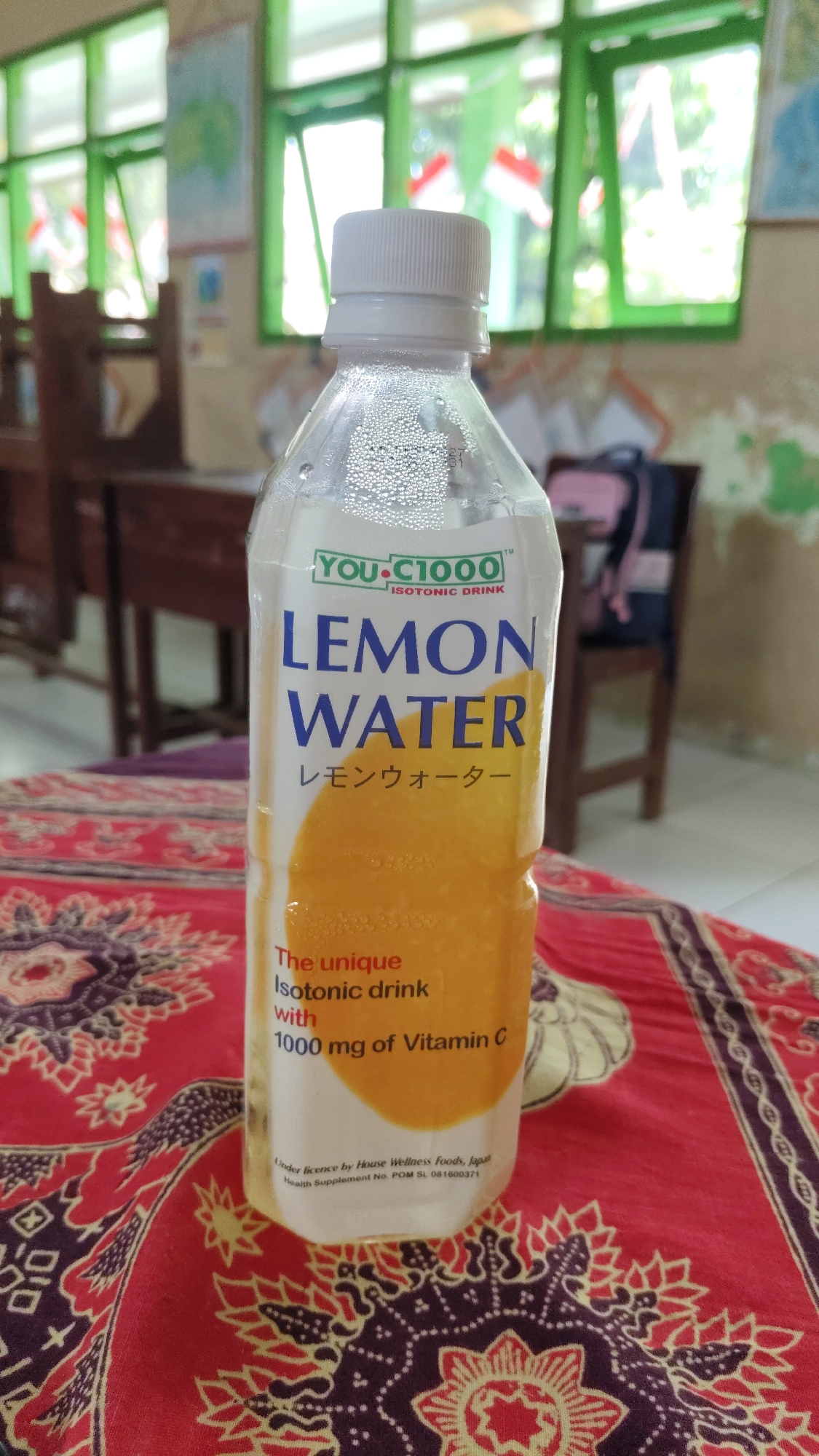 The image shows a close-up of a bottle of "YOU.C1000 LEMON WATER" isotonic drink. The bottle is clear plastic with a white cap and condensation visible on the outside. The label is white with blue and green text, featuring the product name and a graphic of a lemon. Below the main text, there are Japanese characters and smaller English text stating "The unique Isotonic drink with 1000 mg of Vitamin C". At the bottom of the label, there is smaller text indicating "Under licence by House Wellness Foods, Japan" and "Health Supplement No. POM SL 081600371". The bottle is placed on a red fabric with an intricate dark purple and yellow pattern. In the blurred background, wooden tables and chairs are visible, suggesting a classroom or study area. Green-framed windows with Indonesian flags hanging behind them are also apparent, which might indicate the setting is a school in Banjarsari, Indonesia, given the presence of the Indonesian flag. There are no people visible in the image.